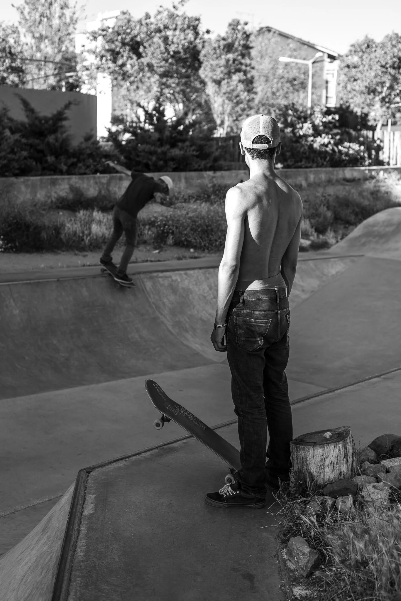 A shirtless young man wearing a cap and jeans standing on a skateboard at a skate park, watching a skateboarder perform tricks on the ramp.