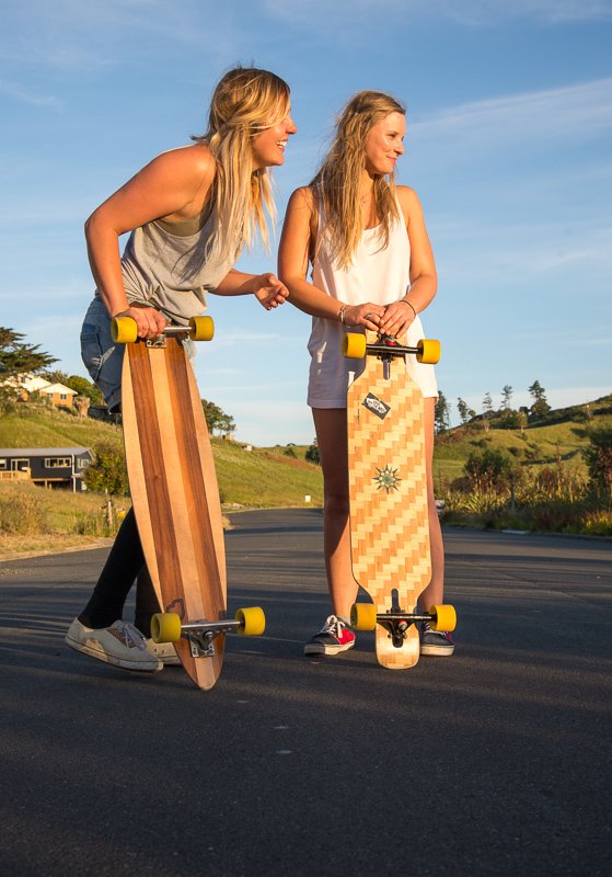 Two women with long blonde hair, wearing casual summer clothes, stand on skateboards on a paved road in a rural area with green hills and houses in the background, smiling and talking to each other during daytime.