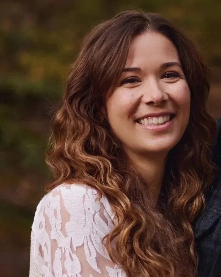 Close-up of a smiling woman with long, curly brown hair, wearing a white lace top, outdoors with a blurred natural background.