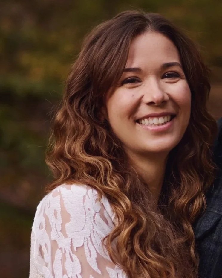 A woman with long, wavy brown hair smiling outdoors with a blurred natural background.