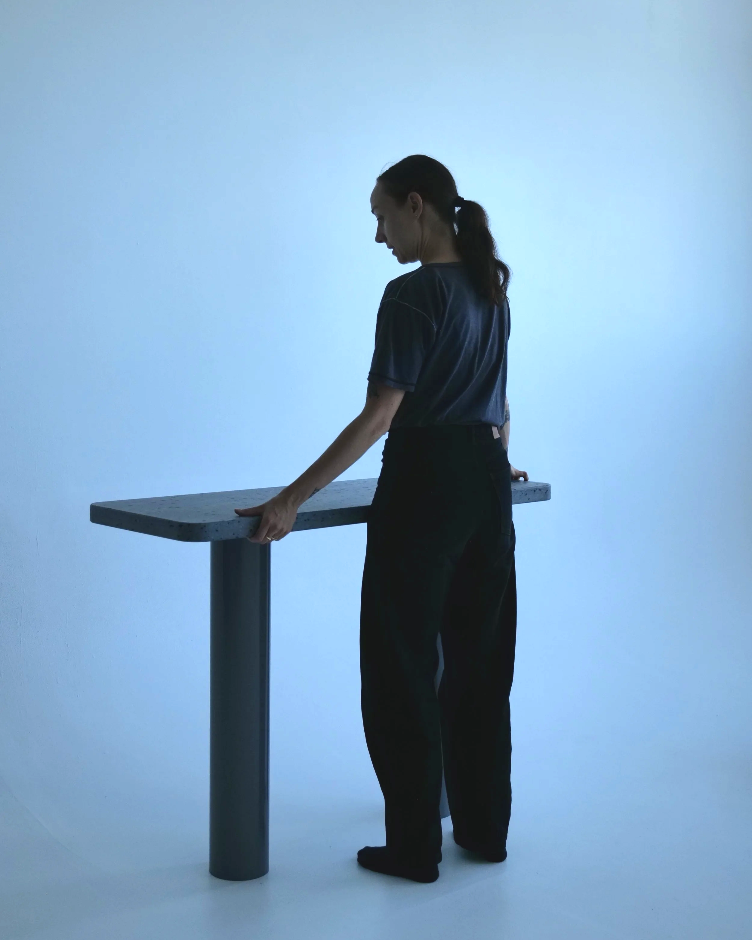 A woman with dark hair tied in a ponytail stands beside a table against a plain light blue background, touching the table with her left hand. She is wearing a dark shirt and pants.