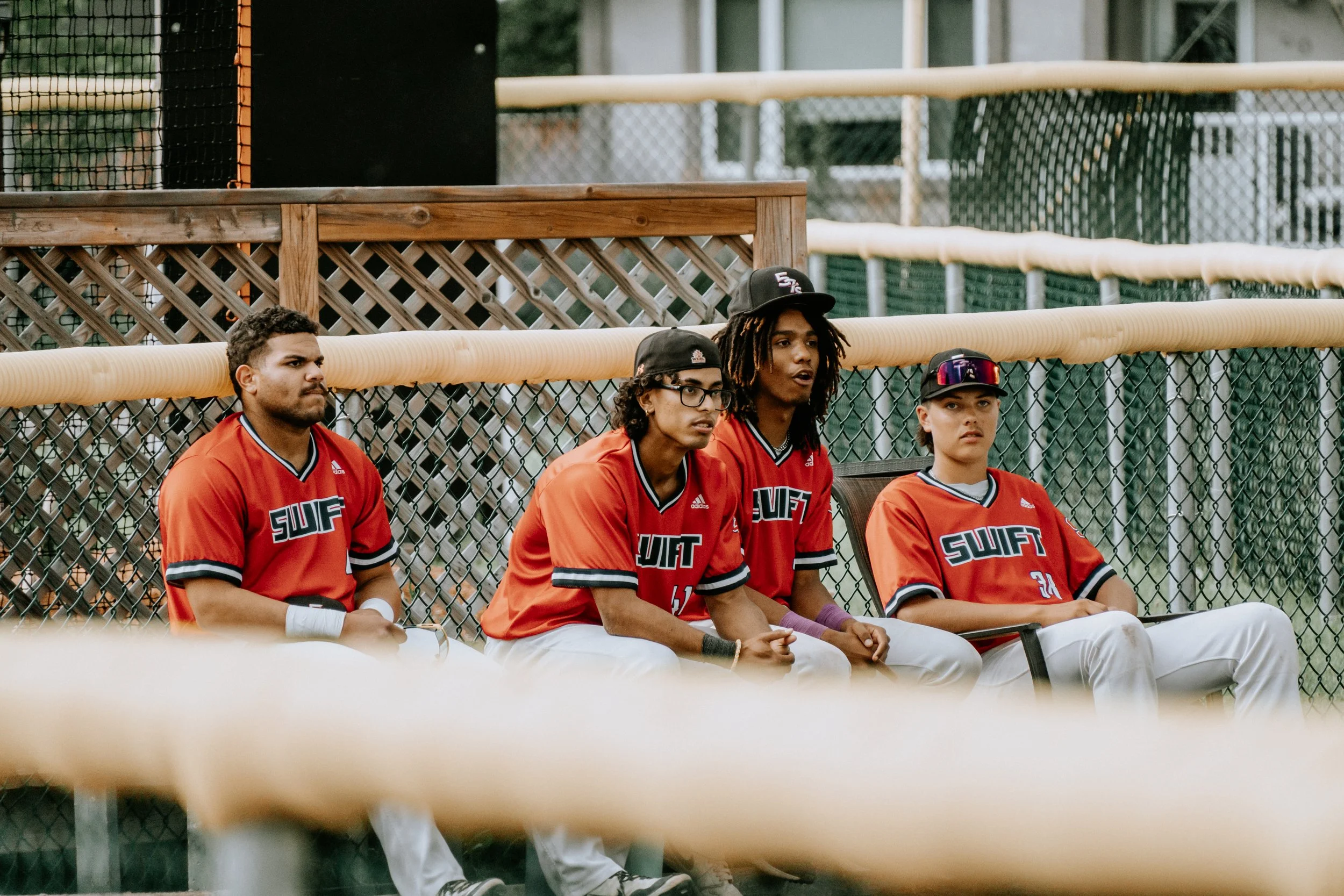 Four young baseball players sitting on a bench in a dugout, wearing red jerseys with the word 'SWIFT' on them. They are behind a chain-link fence, with a wooden partition behind them, at a baseball field.