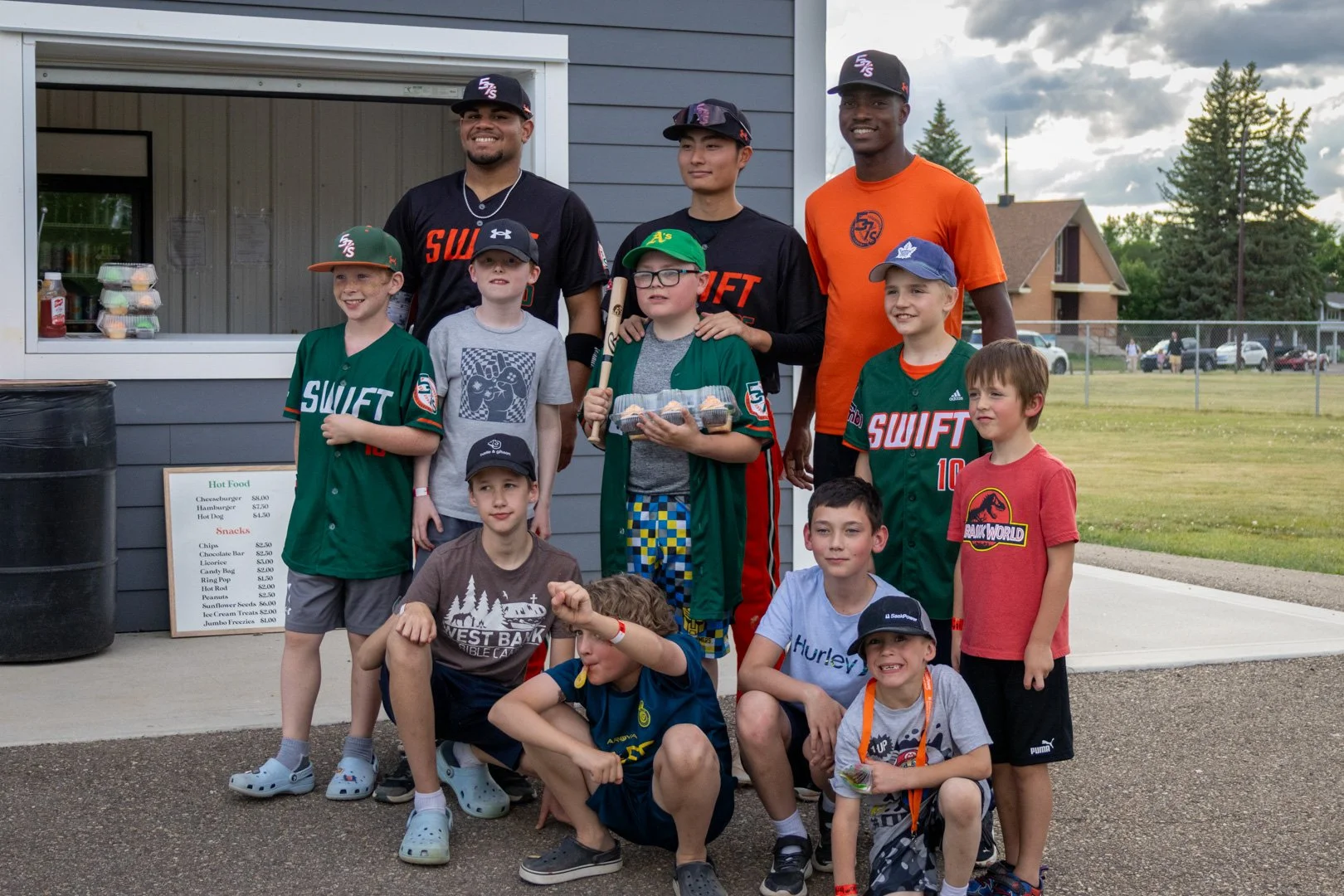 A group of children and adults posing together outdoors in front of a small food stand. The children are wearing various sports jerseys, hats, and casual clothes, some holding snacks or drinks. The adults are dressed in athletic shirts and caps, standing behind the children. There are houses, trees, and a fence in the background.