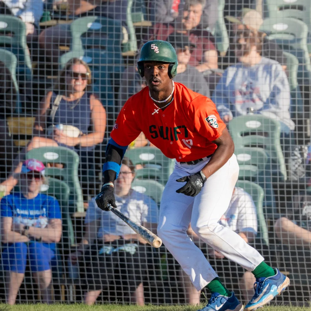A baseball player wearing a red uniform, helmet, and gloves is standing in a batting stance during a game, with an audience in the background.