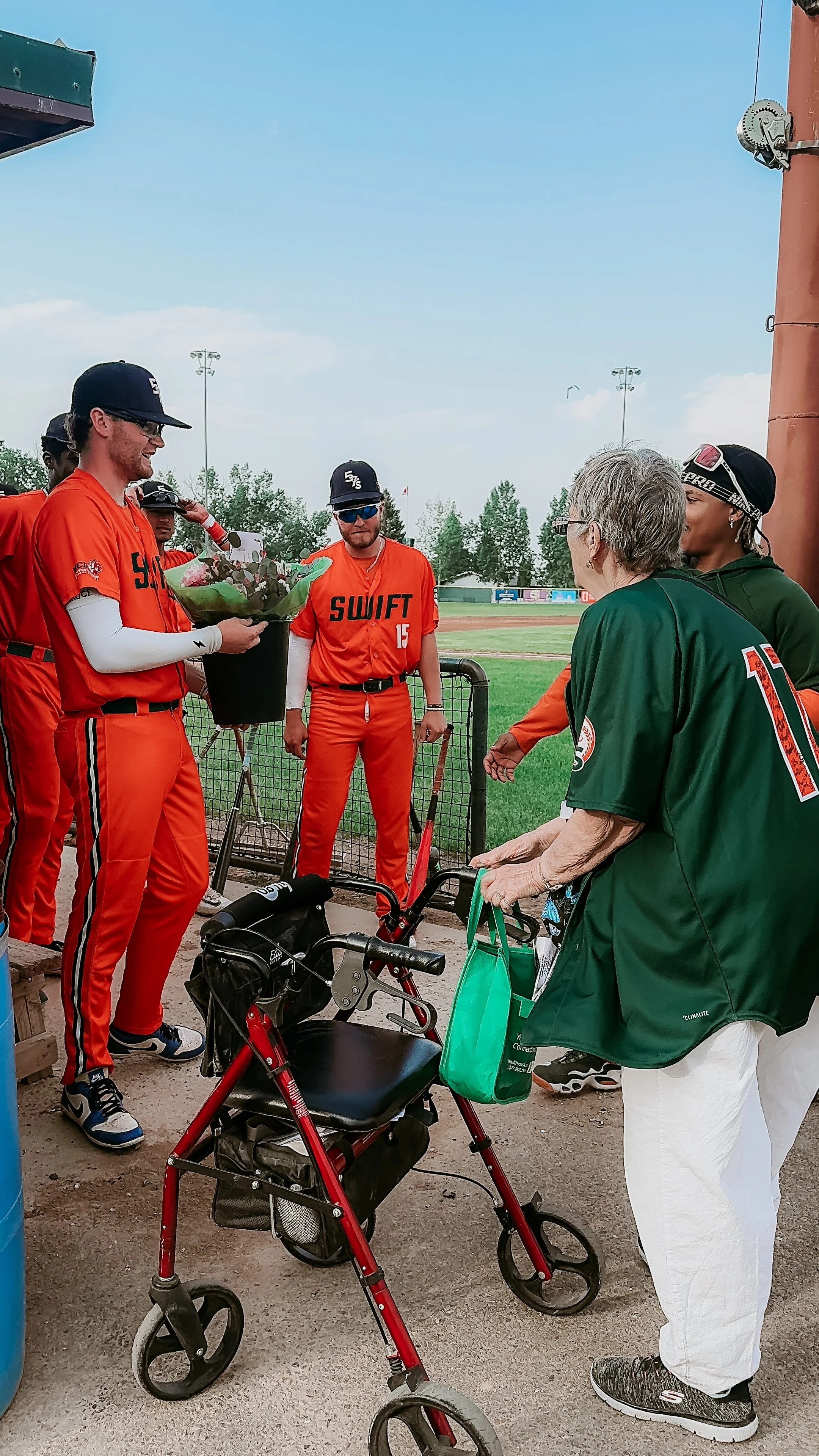 Baseball players in orange uniforms standing near a woman with gray hair and a walker at a baseball field.