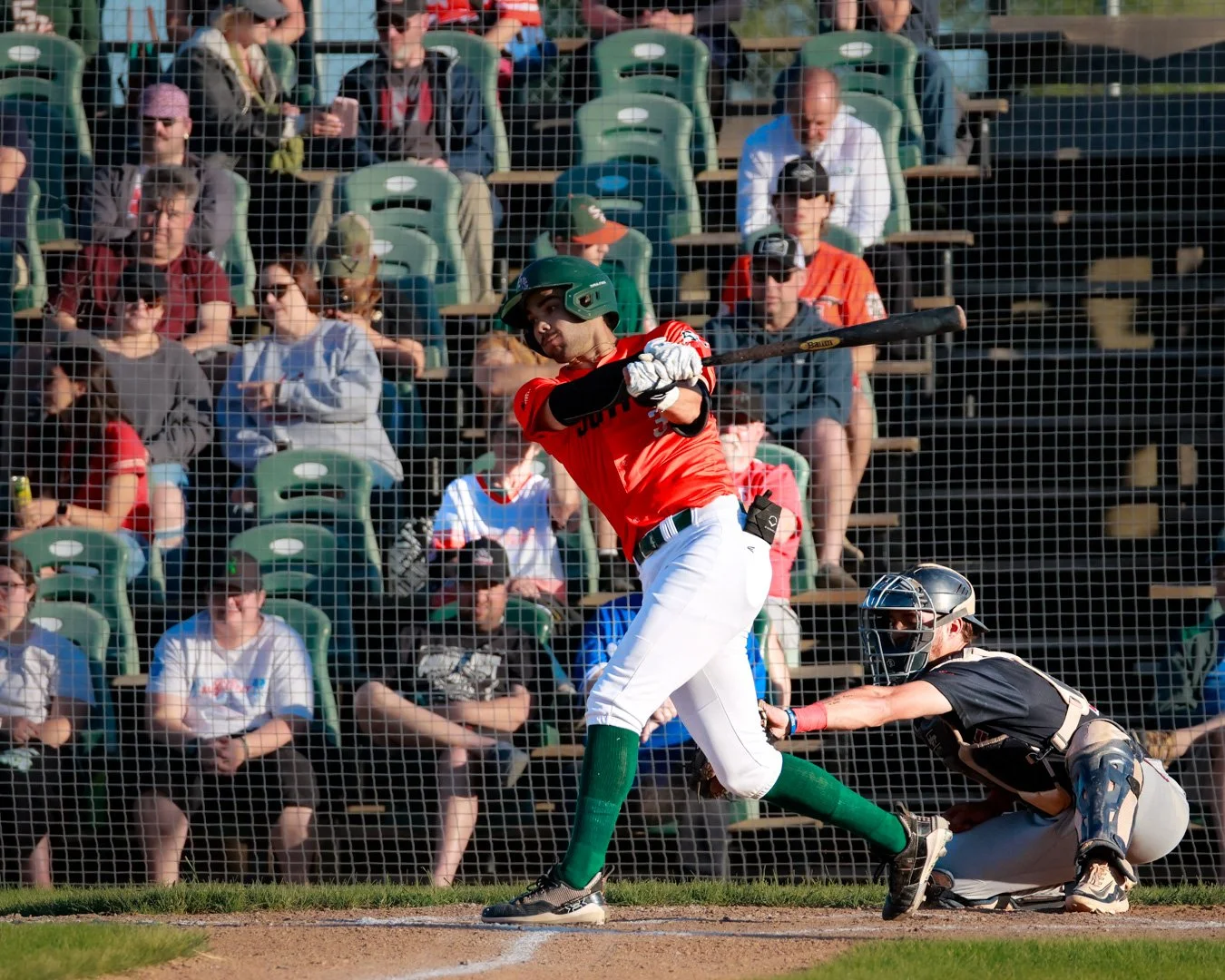 A baseball player in a red uniform and green helmet is swinging a bat at a pitched ball while the catcher in black gear crouches behind home plate. Spectators sit behind a protective net in the background.