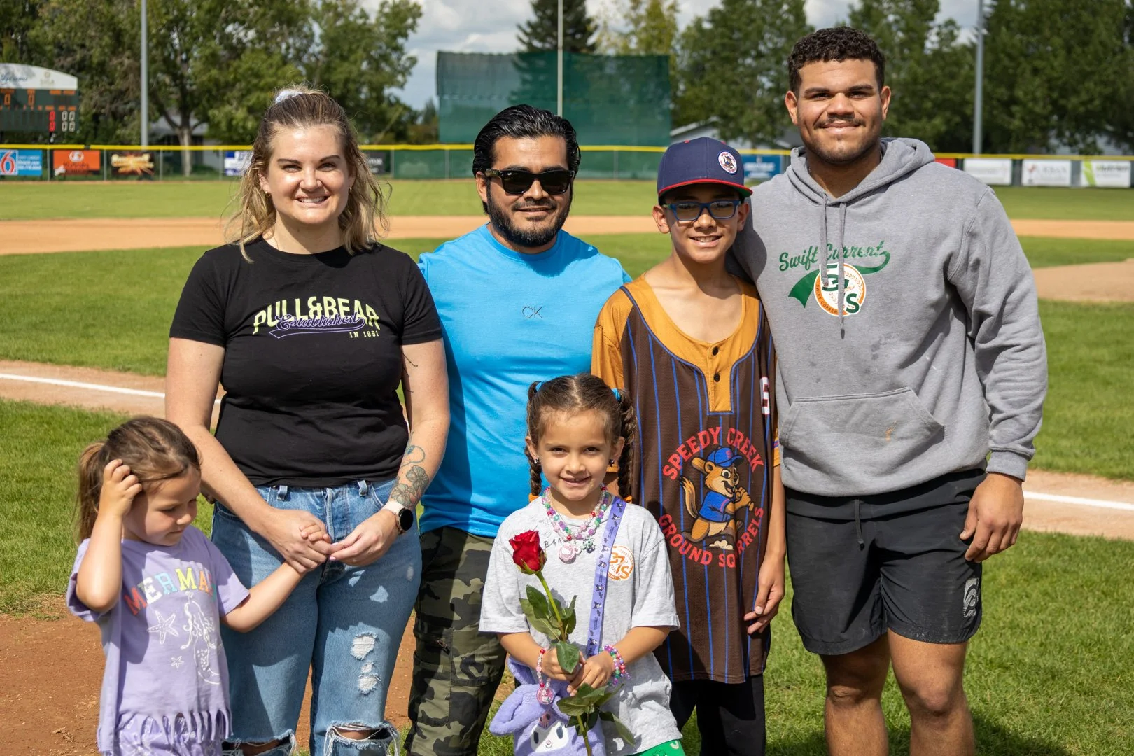 A group of seven people, including children and adults, standing on a baseball field with grass and a scoreboard in the background. The group is smiling and posing for the photo.