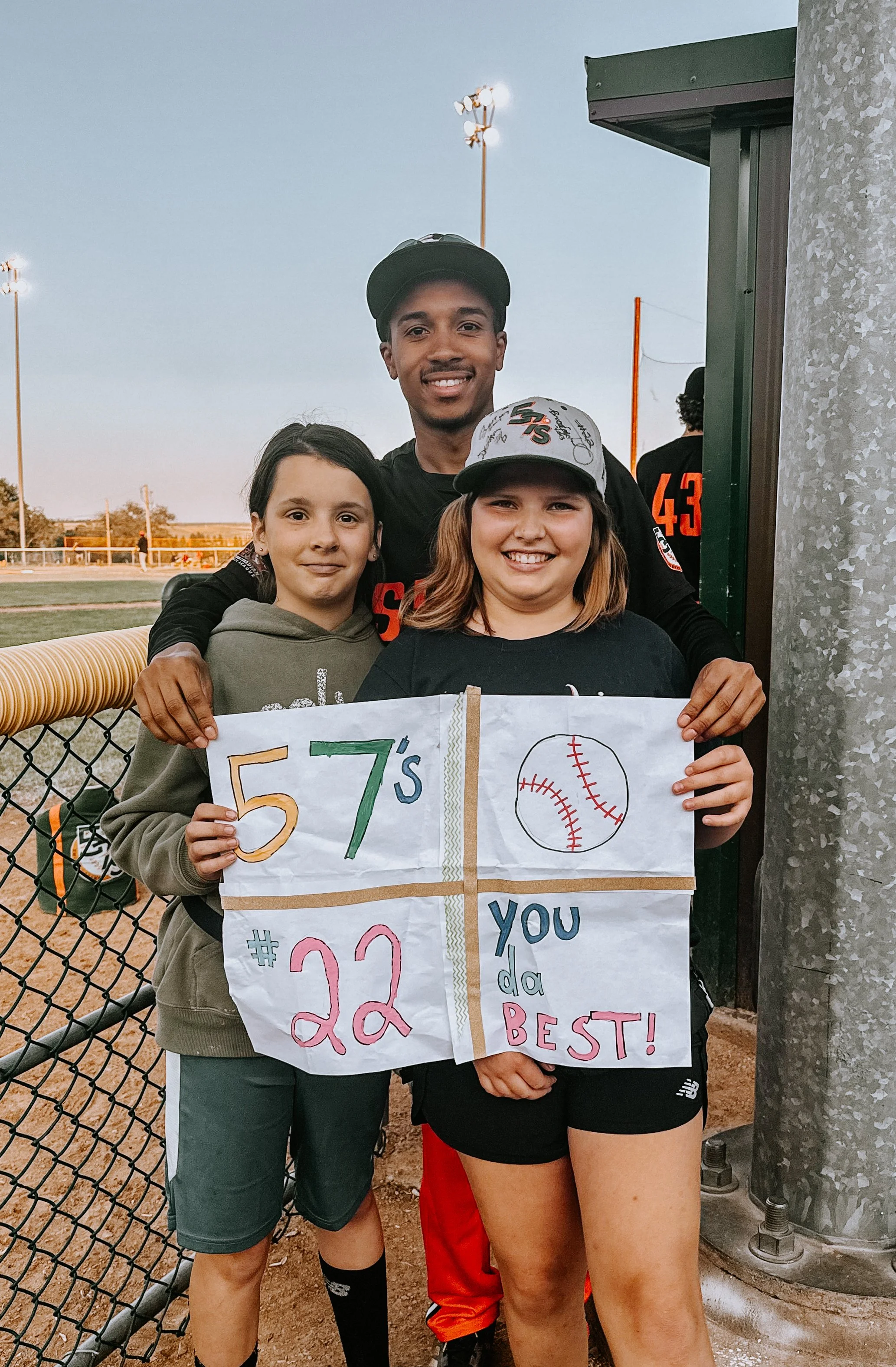 Three young girls and a young man standing by a baseball field fence in the early evening, holding a colorful handmade sign that reads '557's #22 you da best!' with a baseball illustration, wearing athletic clothing and smiling.