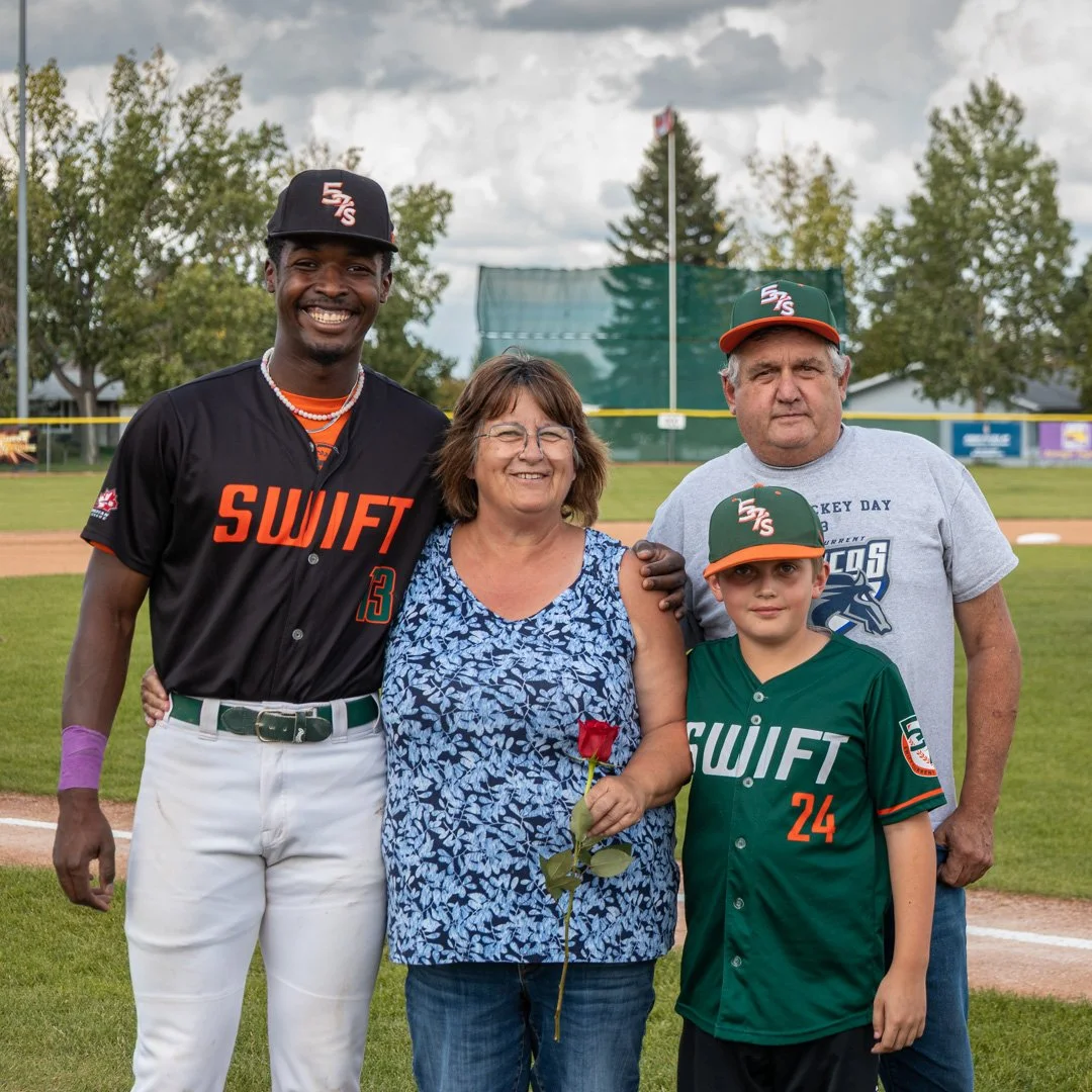 A group of five people standing on a baseball field, smiling at the camera. The group includes a young African American baseball player in a black and orange uniform, an older woman holding a rose, a young boy in a green baseball uniform, and an older man in a gray t-shirt. The background shows a baseball field with trees and cloudy sky.
