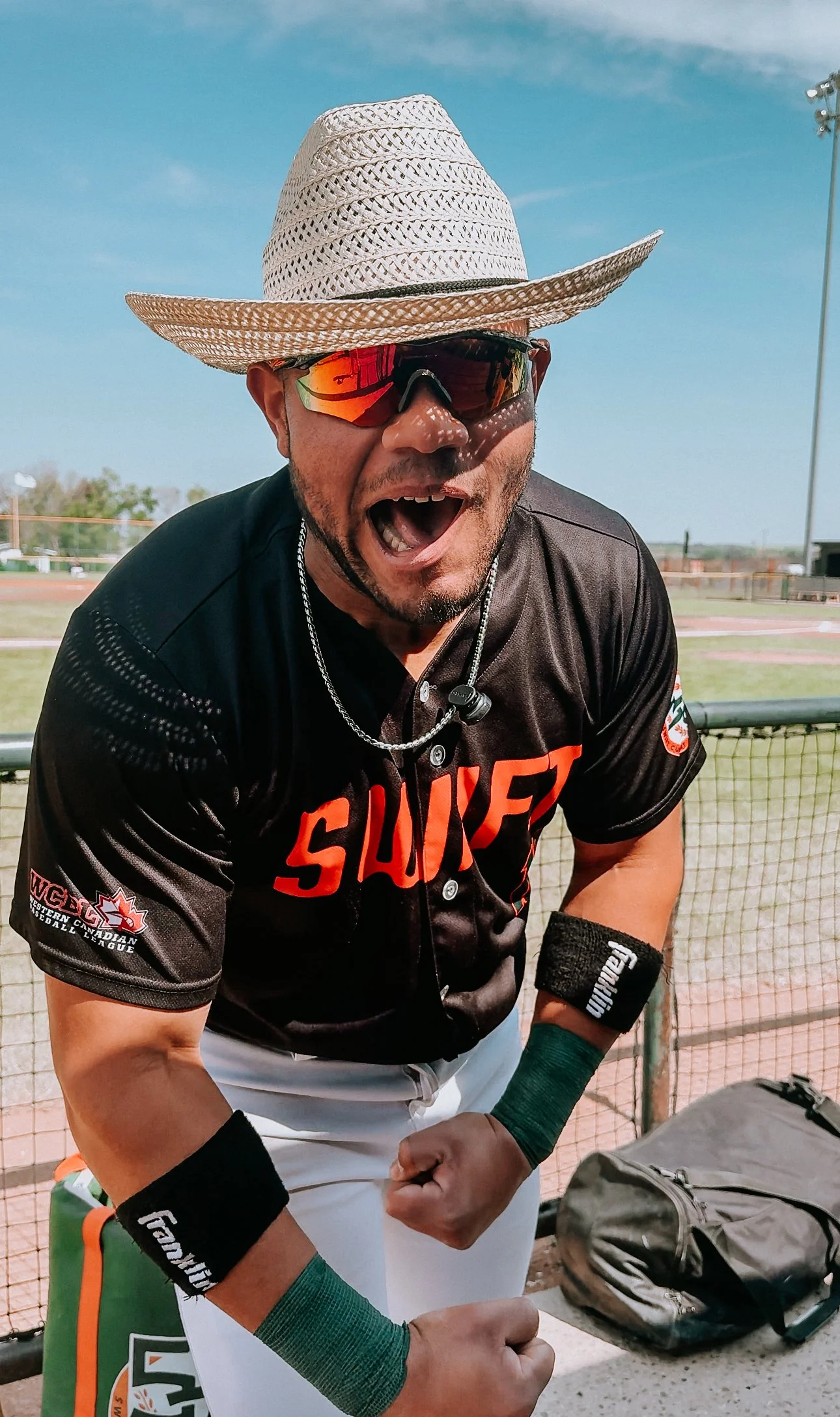 A man wearing sports sunglasses, a wide-brimmed straw hat, and a black sports jersey with red lettering posing with a flexed arm at an outdoor sports field.