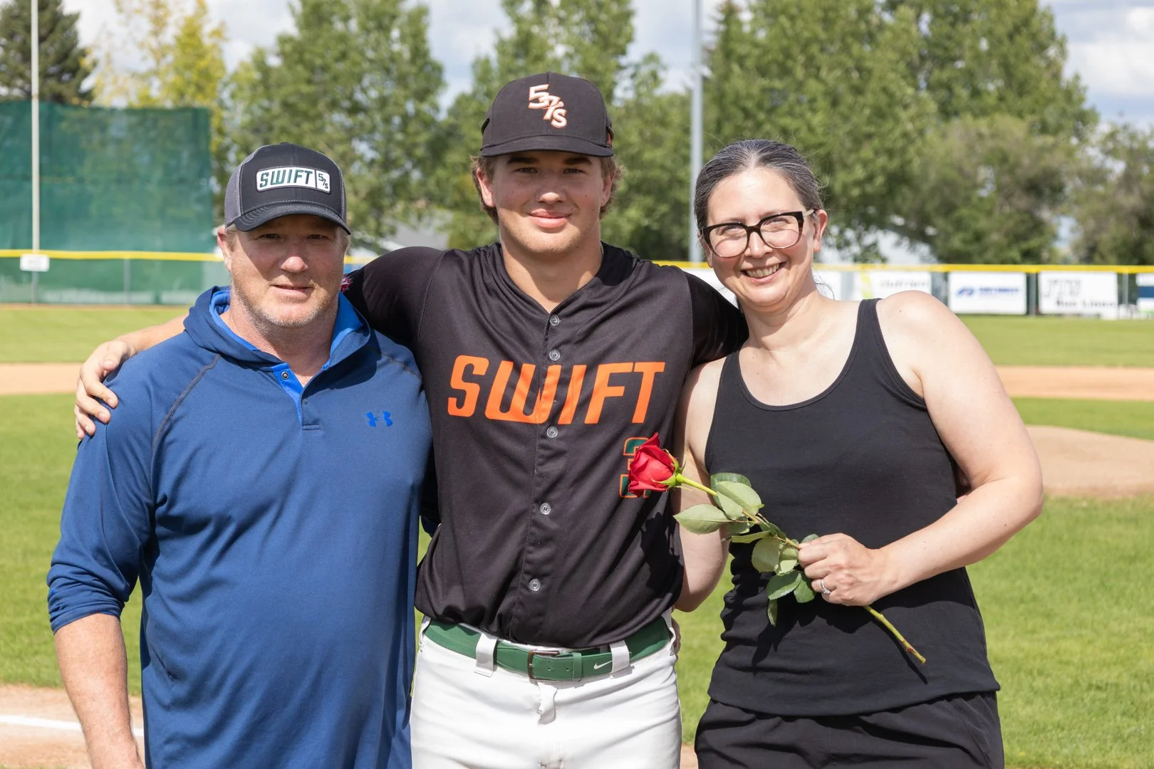 A young male baseball player standing on a baseball field with an older man and woman, all smiling. The woman is holding a red rose.