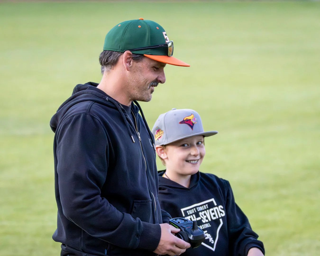 A man and boy standing outdoors on a grassy field. The man is wearing a green and orange cap, black hoodie, and sunglasses, holding a remote control. The boy is wearing a gray cap with a colorful bird logo, a black hoodie with text, and smiling at the camera.