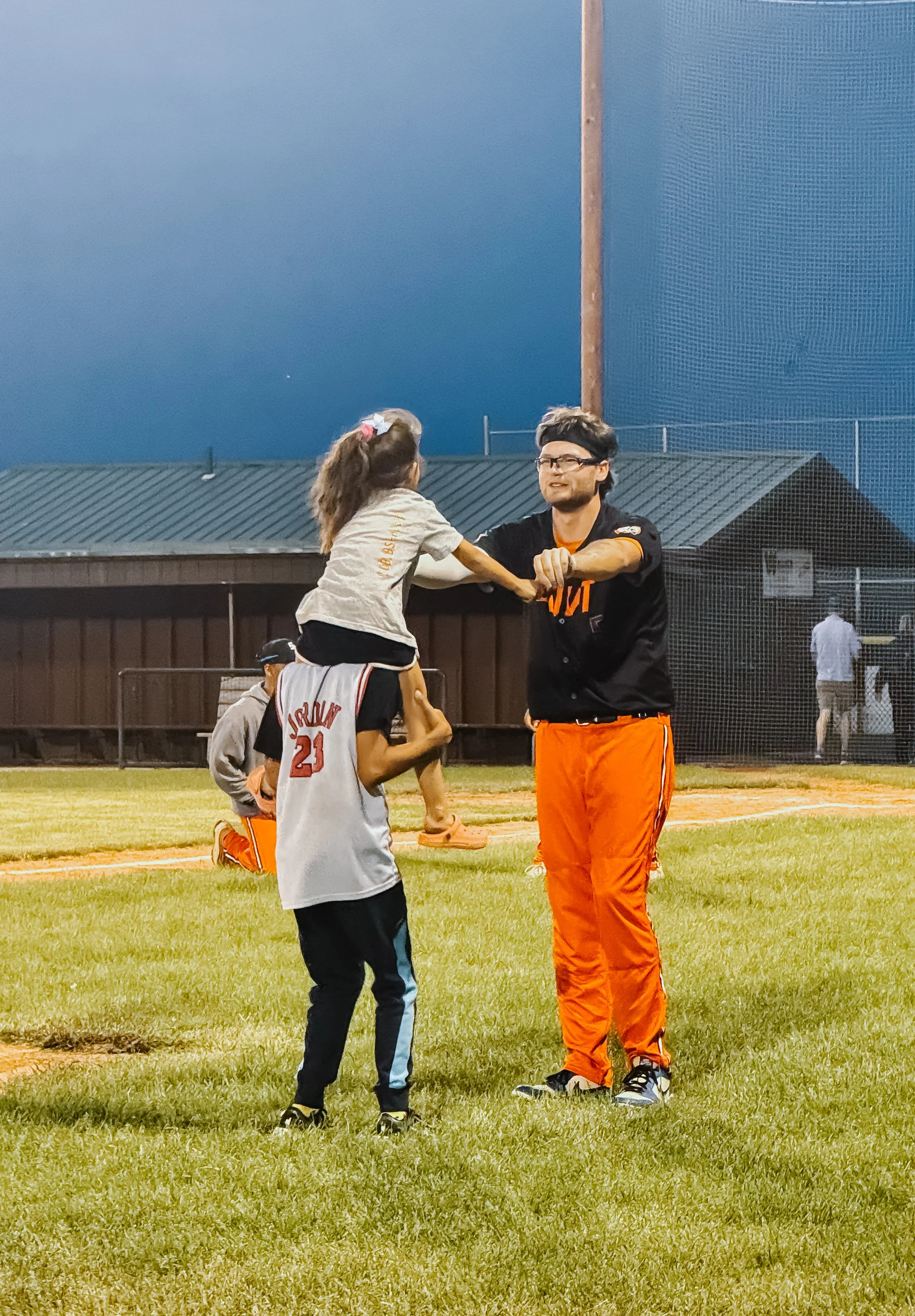 A man and a girl on a baseball field during evening play, the man is in Cleveland Browns gear, and the girl is in a sports uniform with a jersey number 21, holding a baseball bat, with other children and a coach in the background.