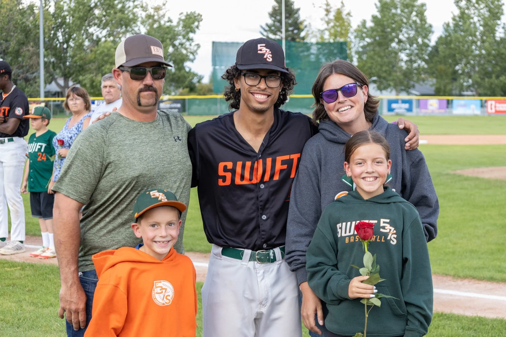 A group of five people, including a young man wearing a black SWIFT jersey, a woman, a man, and two children, standing on a baseball field, smiling for a photo. The children are holding a red rose, and the background shows other players and spectators.