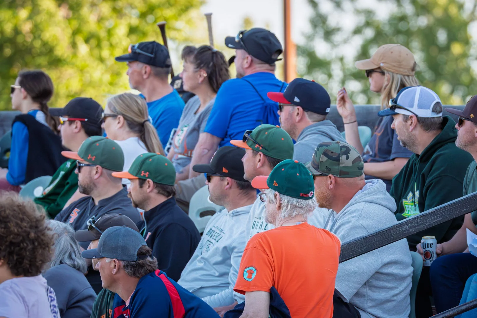 A diverse group of people sitting in outdoor bleachers watching an event, with many wearing baseball caps and casual clothing.