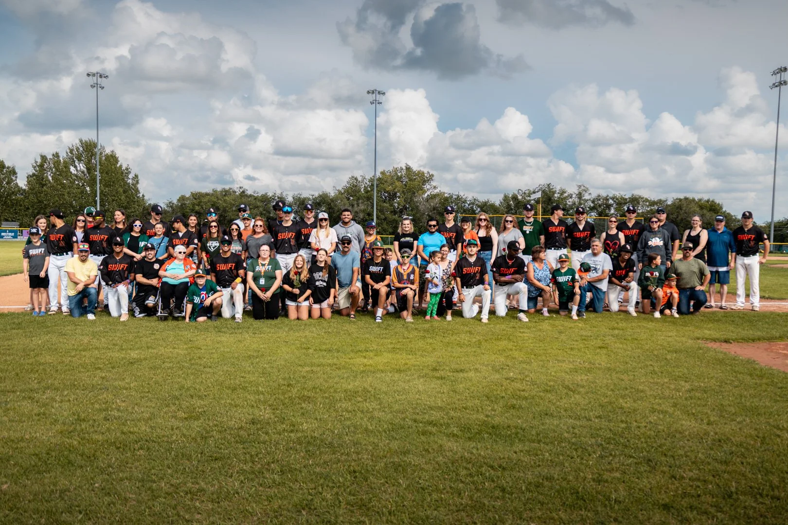 Large group of people, including children and adults, gathered on a baseball field, posing for a group photo. Many are wearing baseball jerseys, caps, and casual clothing. The background shows trees, baseball field lights, and a cloudy sky.