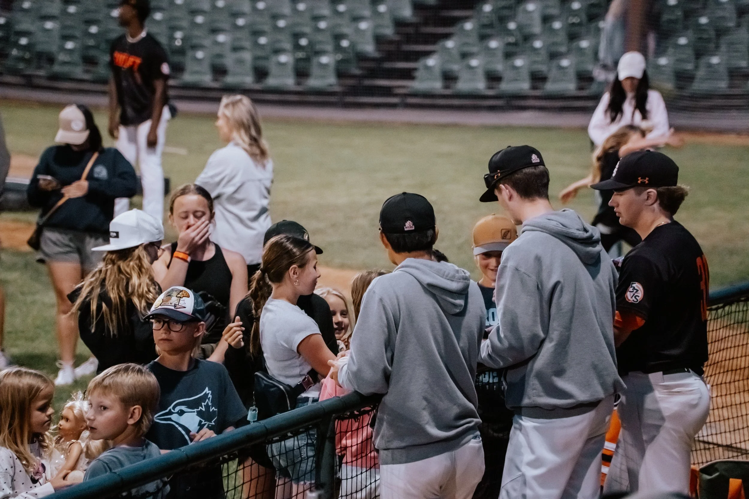 Group of young people, including boys and girls, gathered on a baseball field at night. Some are signing autographs and interacting with fans near the fence, while others stand in the background. The atmosphere appears lively and social.
