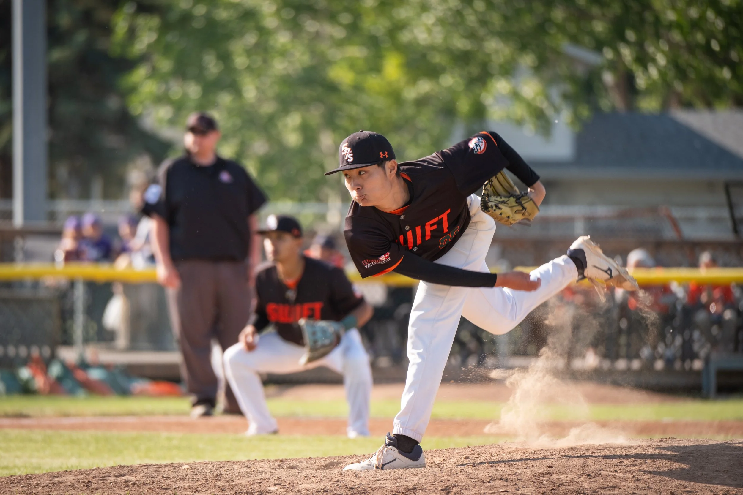 A baseball player in a black and orange uniform is pitching on a baseball field, with teammates and a coach or umpire in the background. The player is in mid-throw, focused and determined.