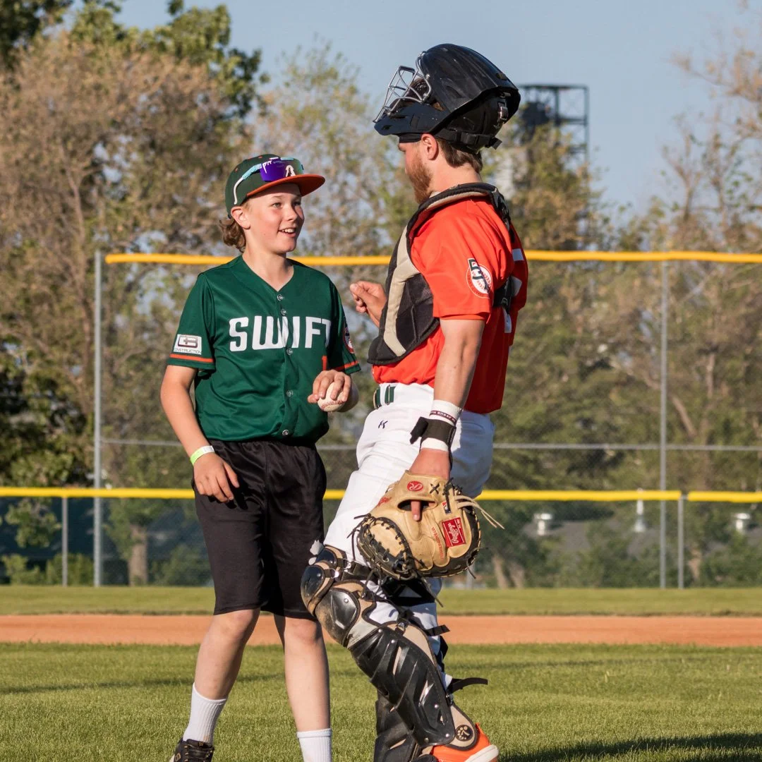 A young baseball player in a green jersey and a baseball cap talking to a coach in a catcher's uniform including a helmet, chest protector, and leg guards, on a baseball field during daytime.