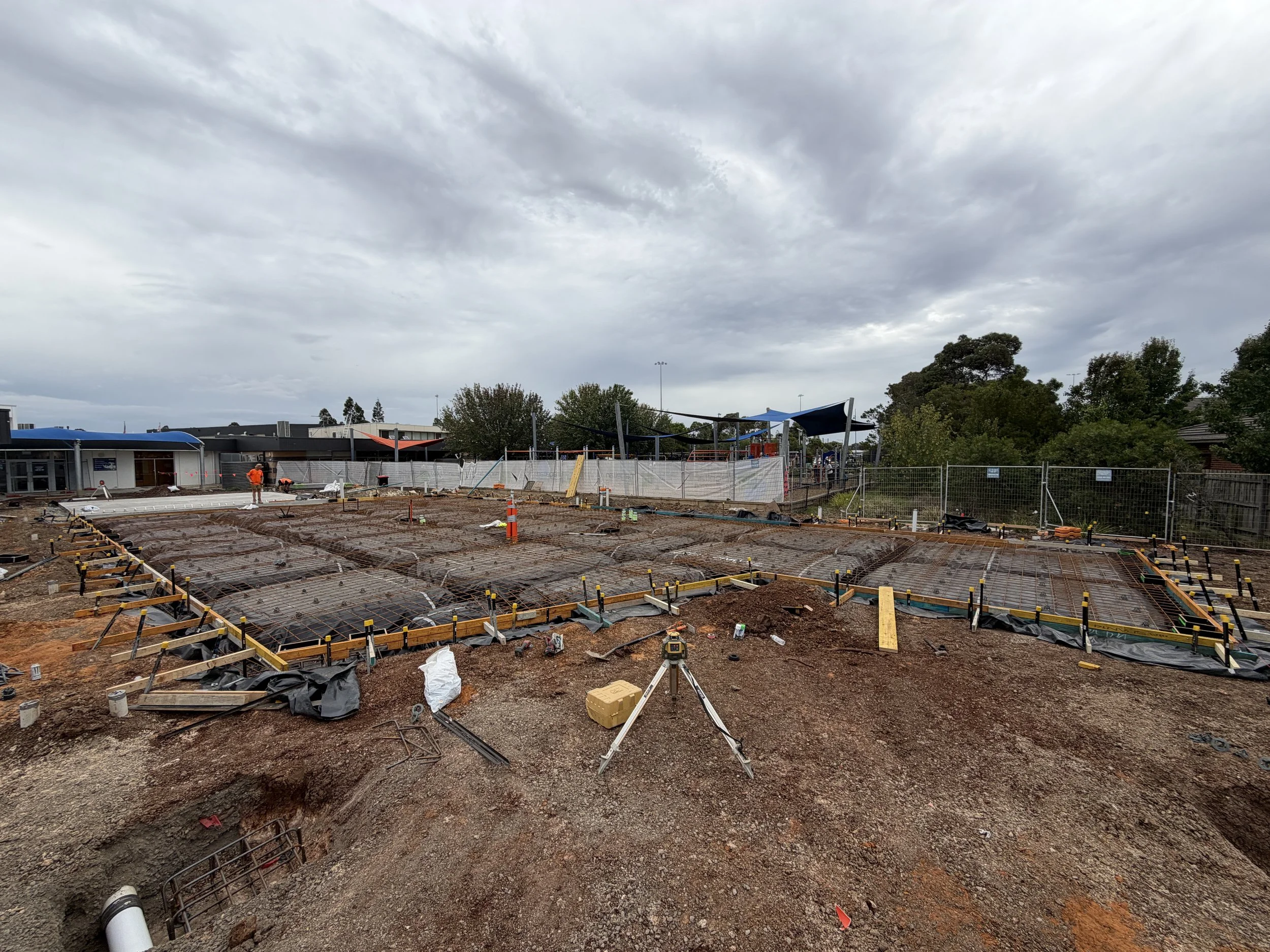 Construction site with workers, partially built foundation, construction materials, and equipment under a cloudy sky.