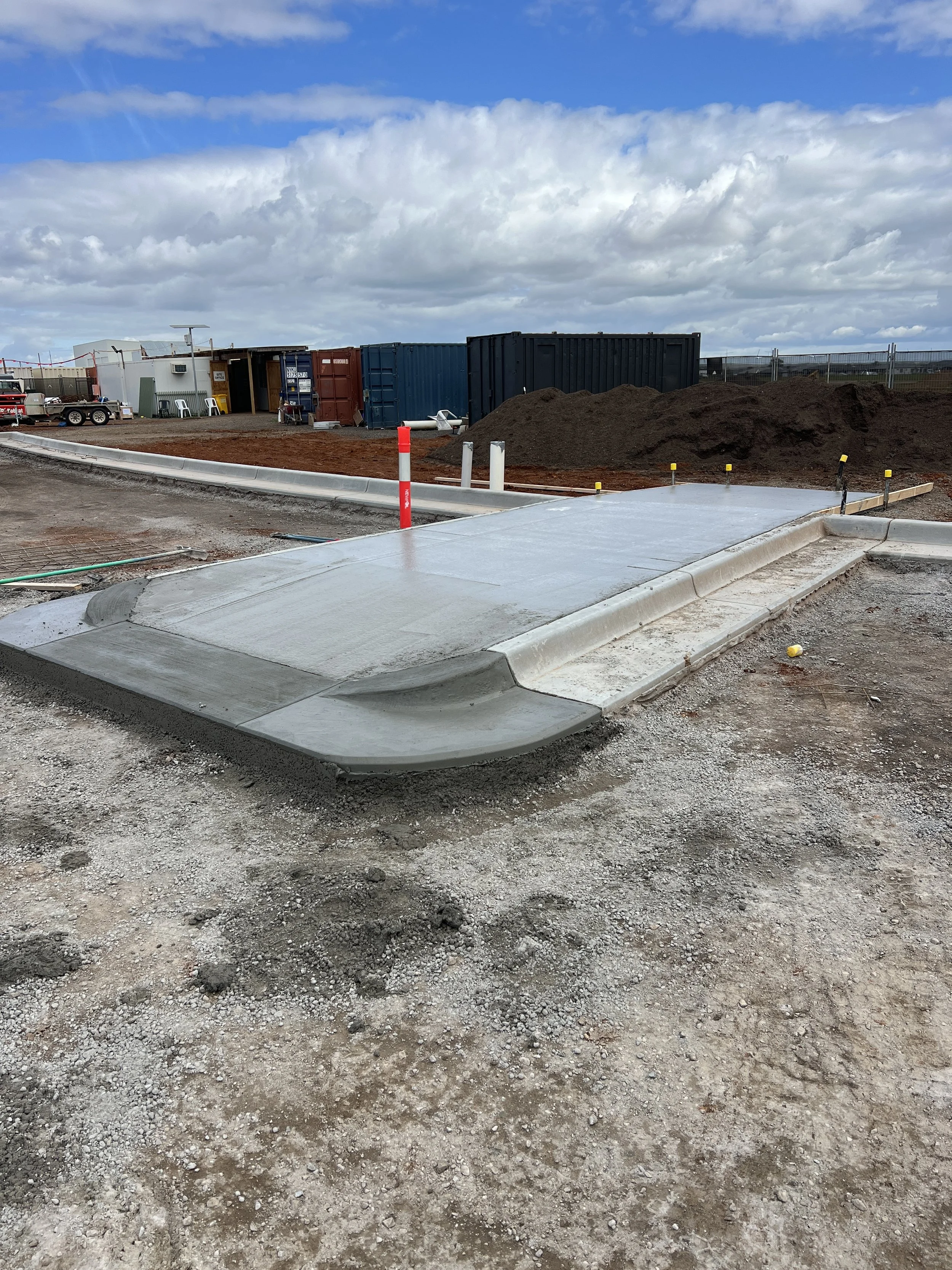 A construction site with freshly poured concrete curbing, gravel, large dirt piles, and storage containers under a partly cloudy sky.
