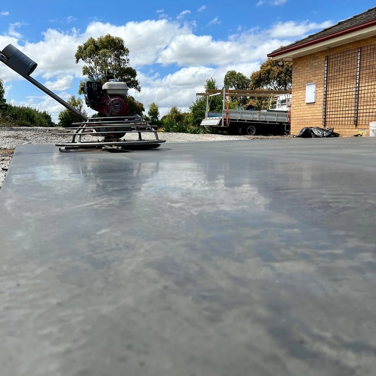Close-up of a concrete surface with a power trowel in the background, outdoors near a brick building under a partly cloudy sky.