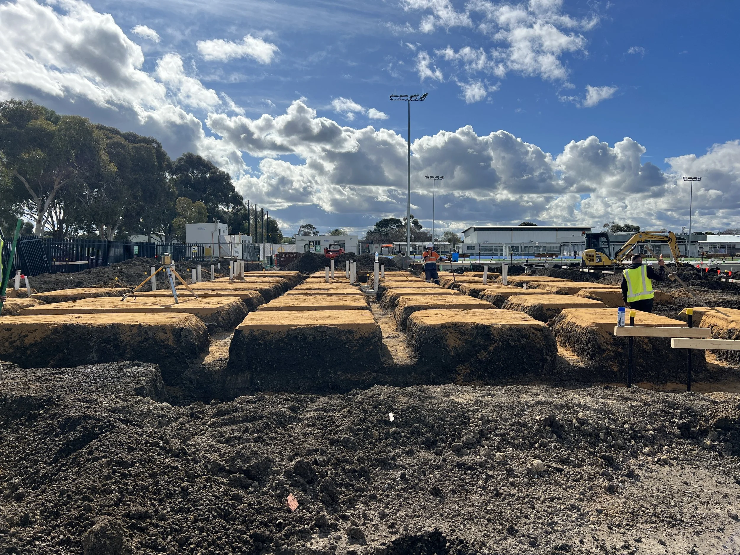 Construction workers are working on an outdoor site with dirt, trenches, and heavy machinery under a partly cloudy sky.
