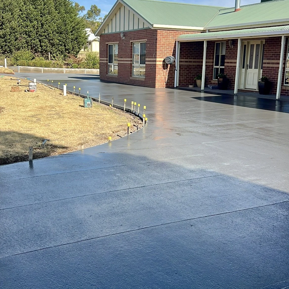 Newly poured concrete driveway in front of a brick house with green roof, surrounded by a partially landscaped yard with yellow stakes and tools nearby.