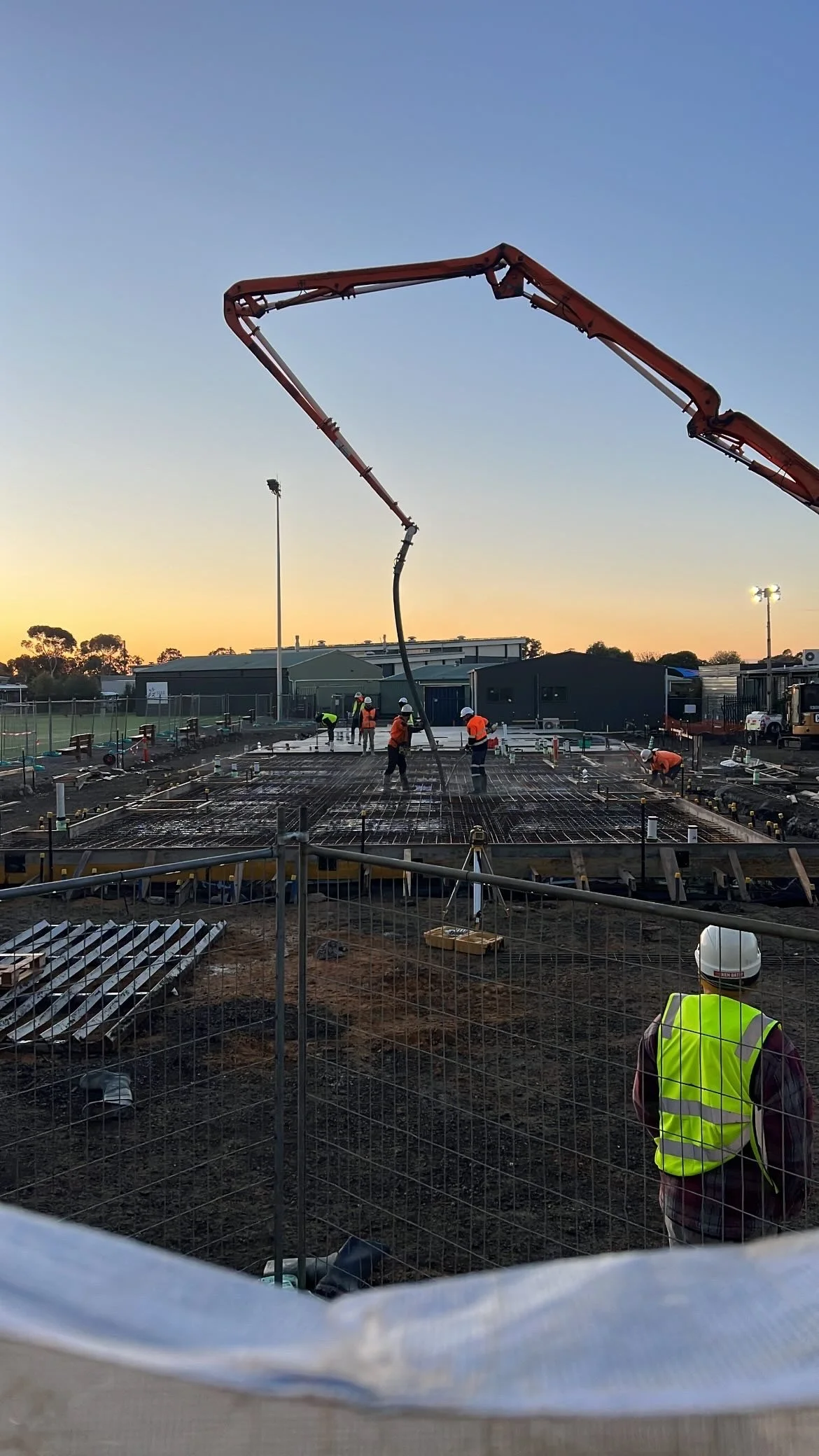Construction workers pouring concrete on a building site during sunset, with cranes and equipment, and safety gear.