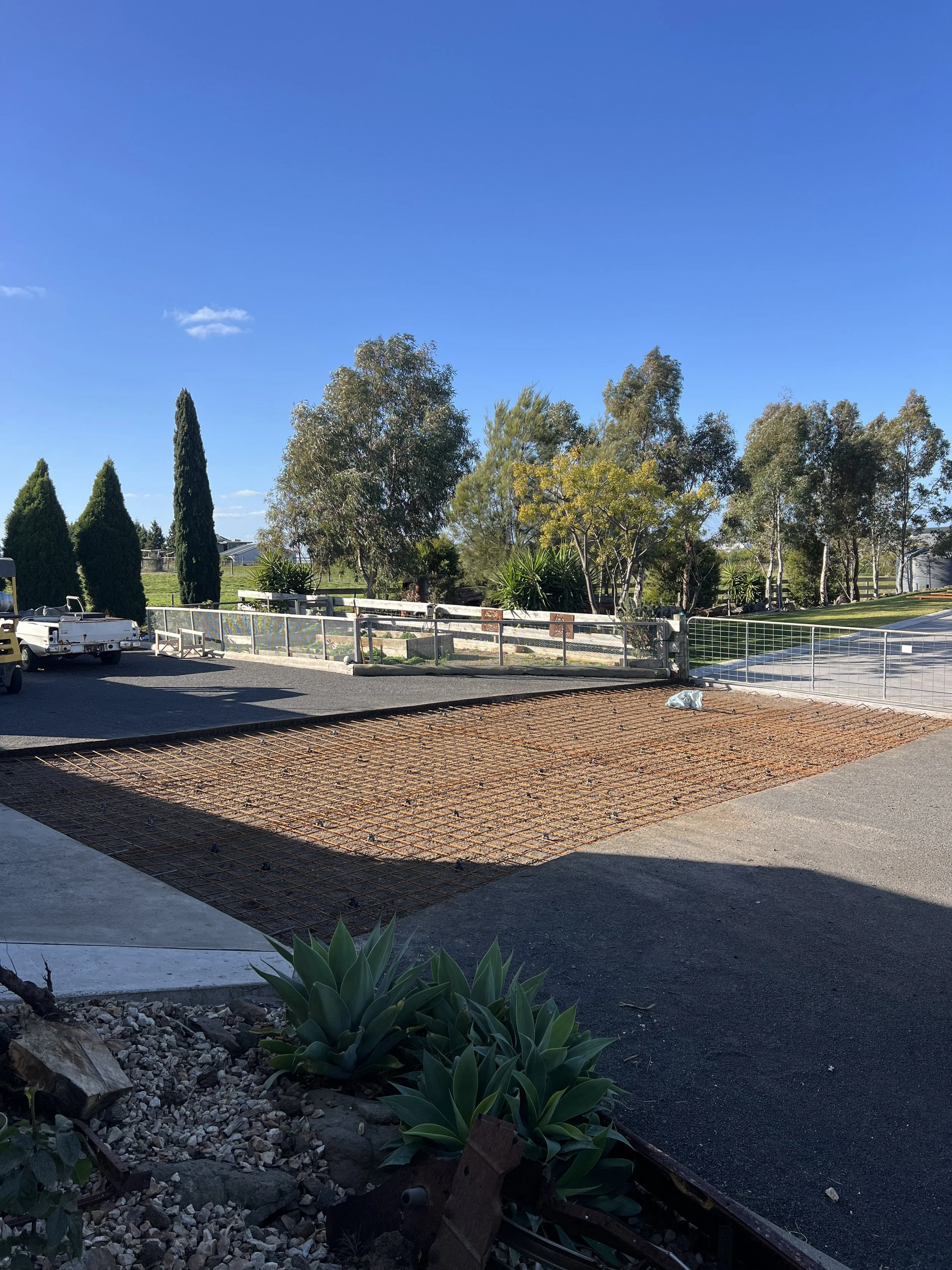 A construction site with a metal grid base, a few plants in the foreground, and trees and a fence in the background under a bright blue sky.