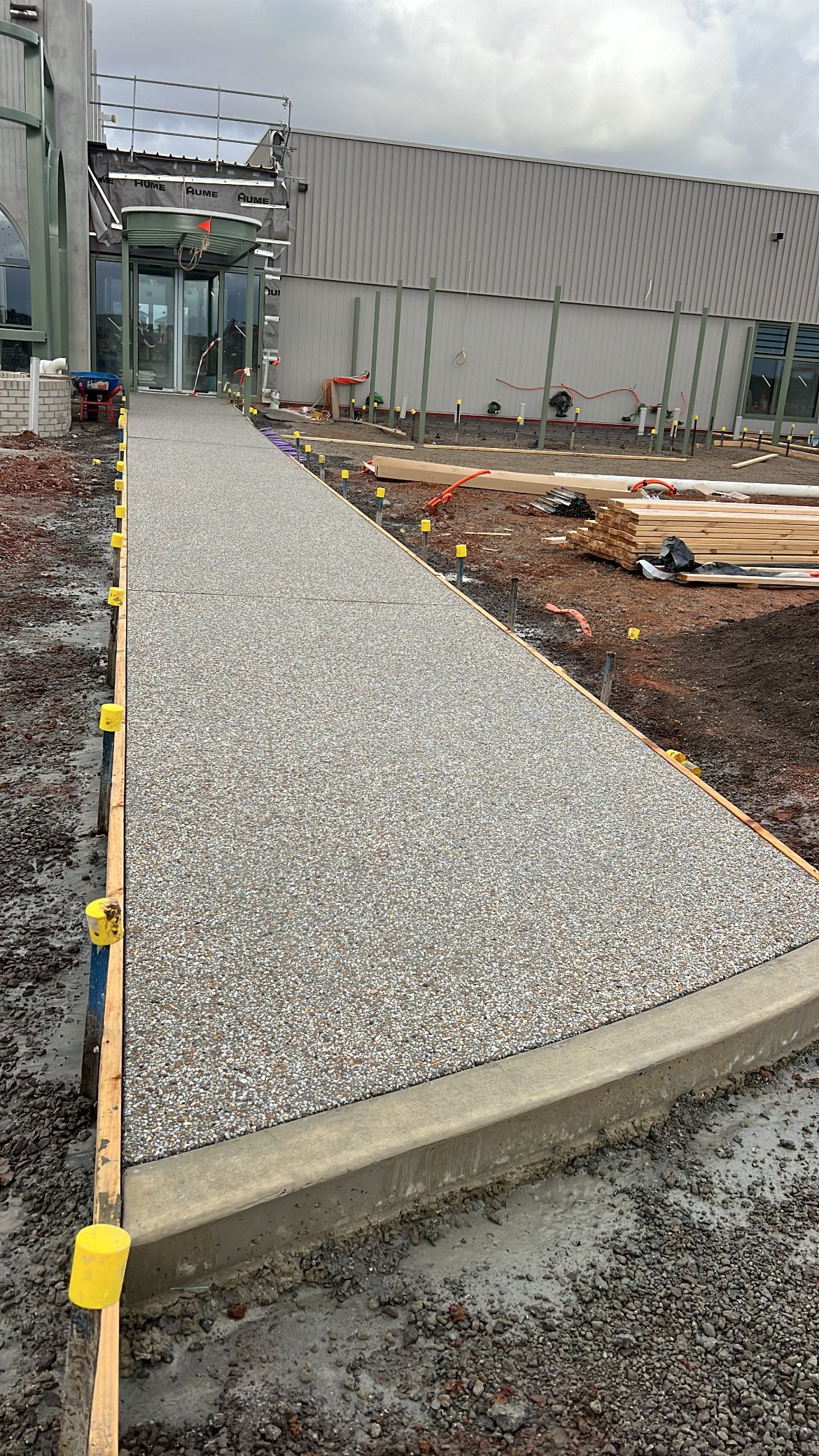 Concrete sidewalk under construction at a building site, with soil, stakes, and building materials around.