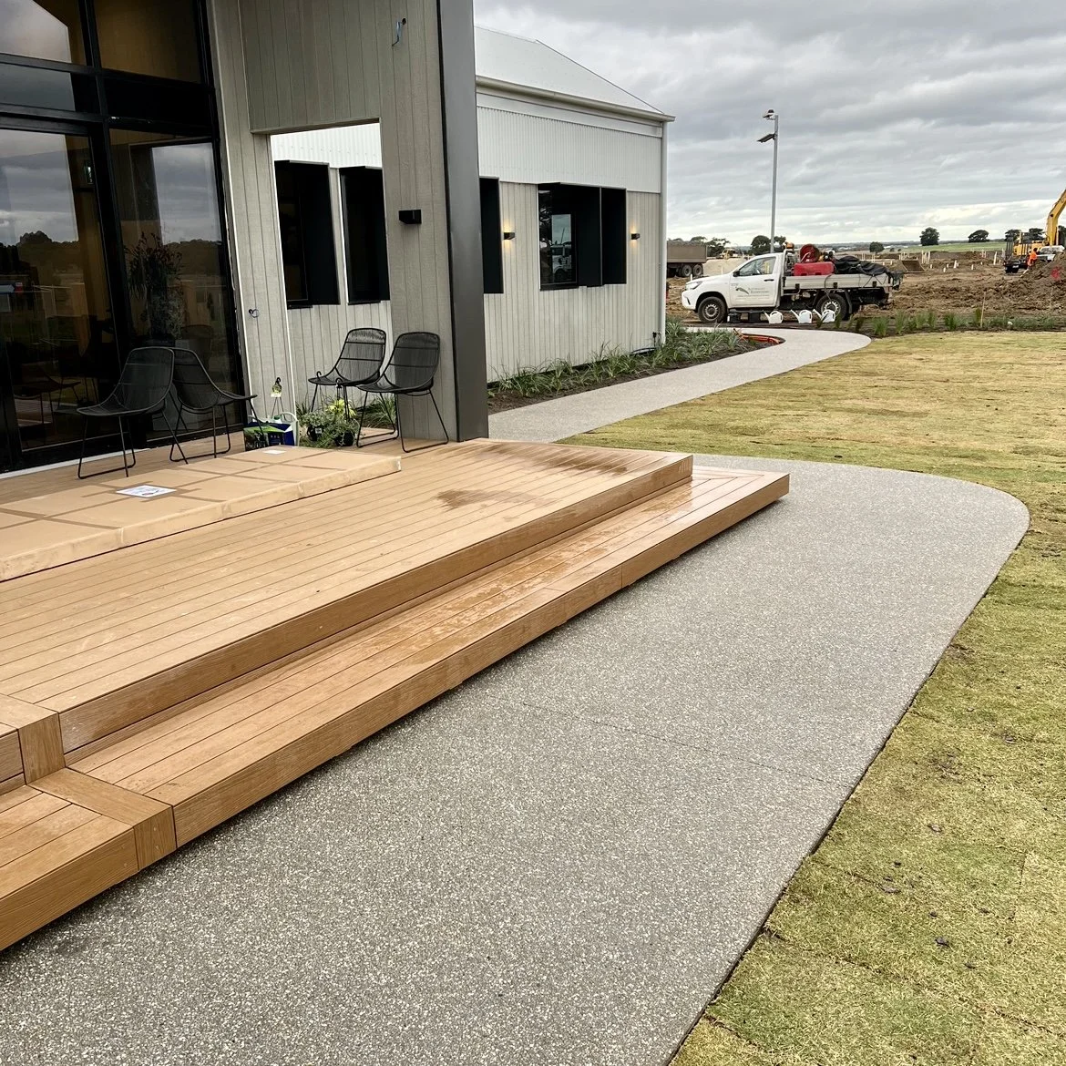 Construction site with a modern building, wooden deck, patio chairs, and a driveway leading to a parking lot with trucks and construction equipment in the background.