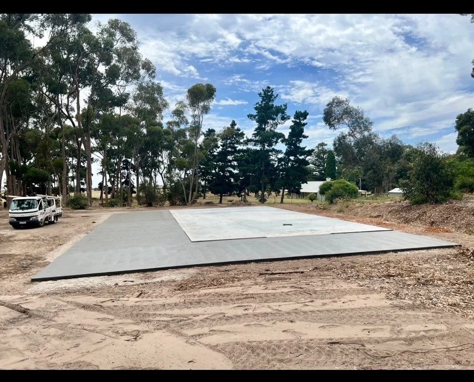 A cleared construction site with a concrete foundation in a rural area, surrounded by trees and a partly cloudy sky.