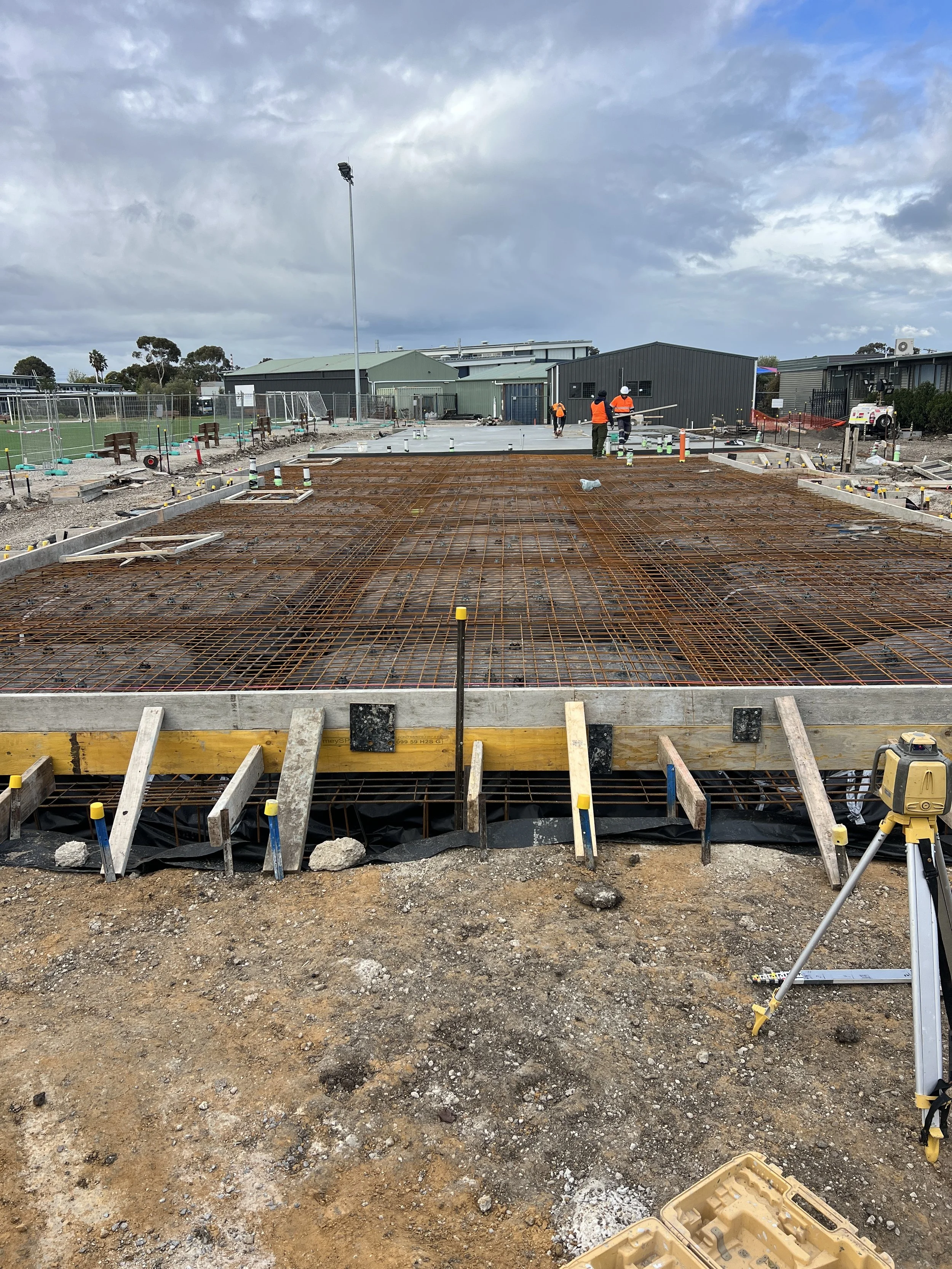 Construction site with workers and rebar for concrete foundation, surrounded by dirt and unfinished structures, under a cloudy sky.
