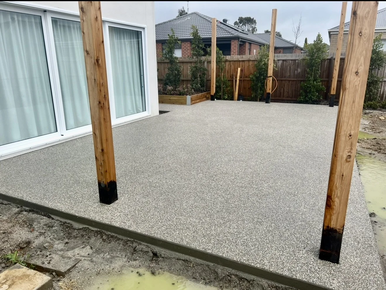 Photo of a newly installed backyard deck with a textured surface, supported by wooden posts, next to a house with large sliding glass doors, and a wooden fence with small trees in the background.