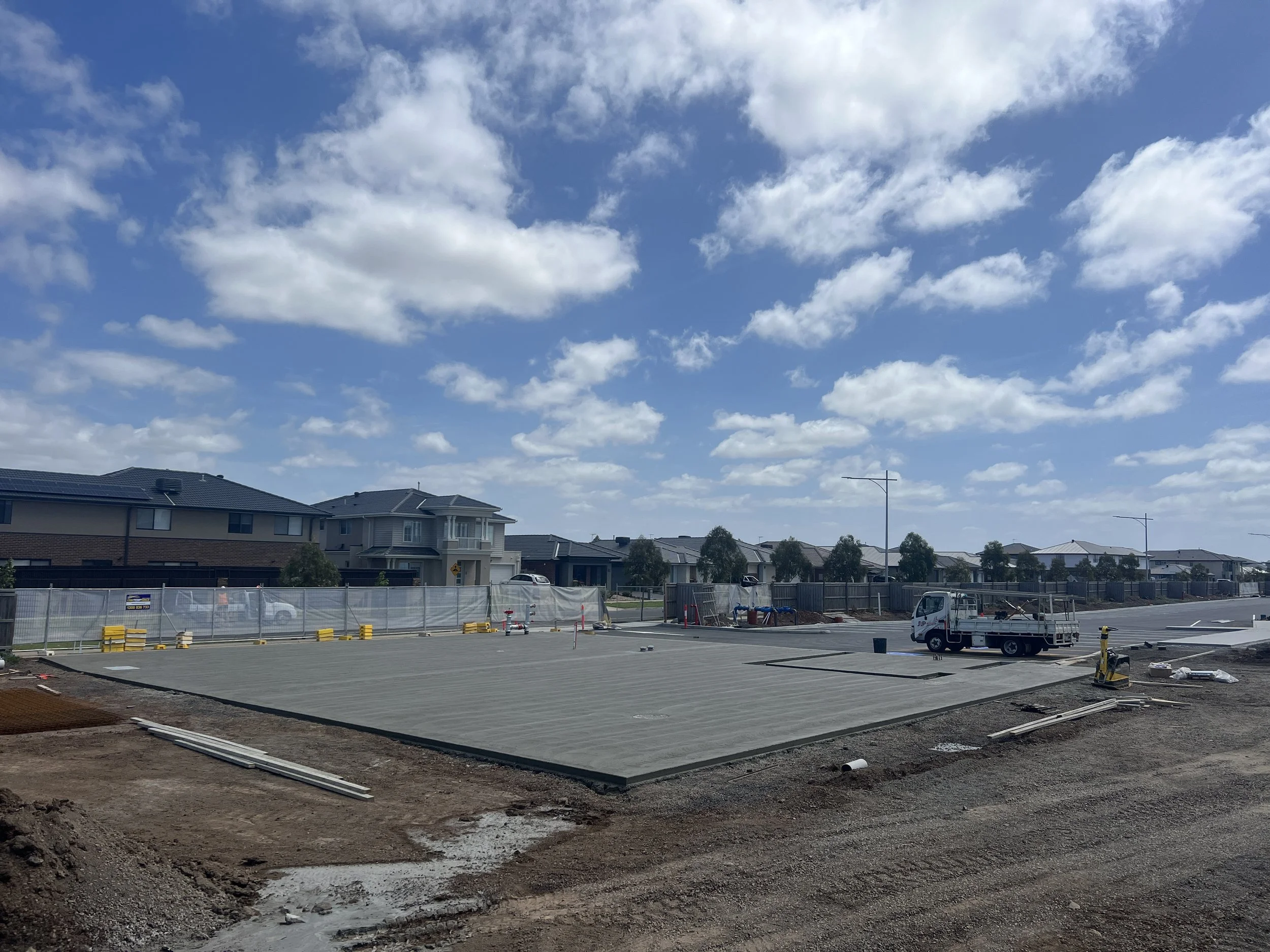 Construction site with a newly poured concrete foundation, construction equipment, and a residential neighborhood with houses under a partly cloudy sky.