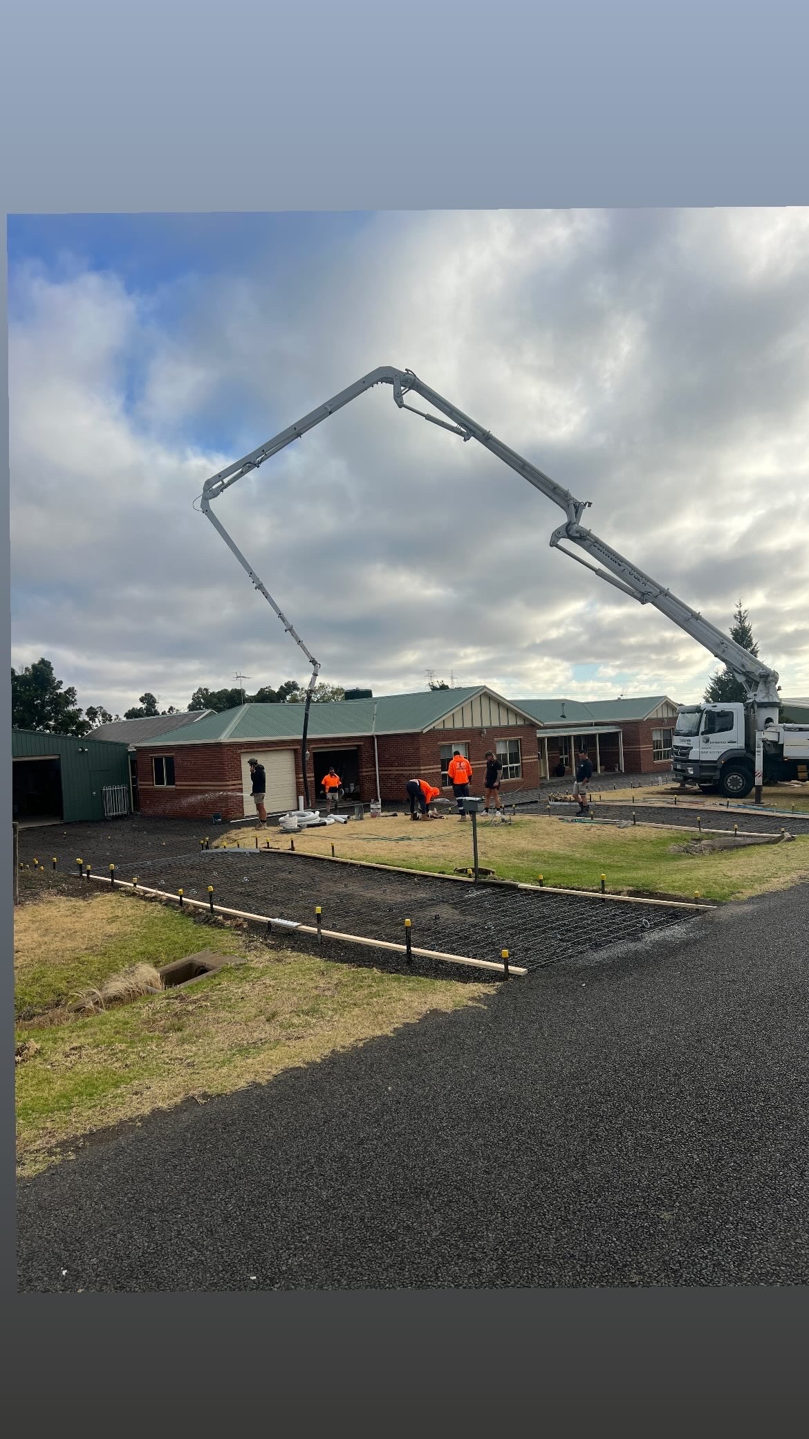Construction workers pouring concrete in front of a house under a cloudy sky, with construction equipment and materials visible.