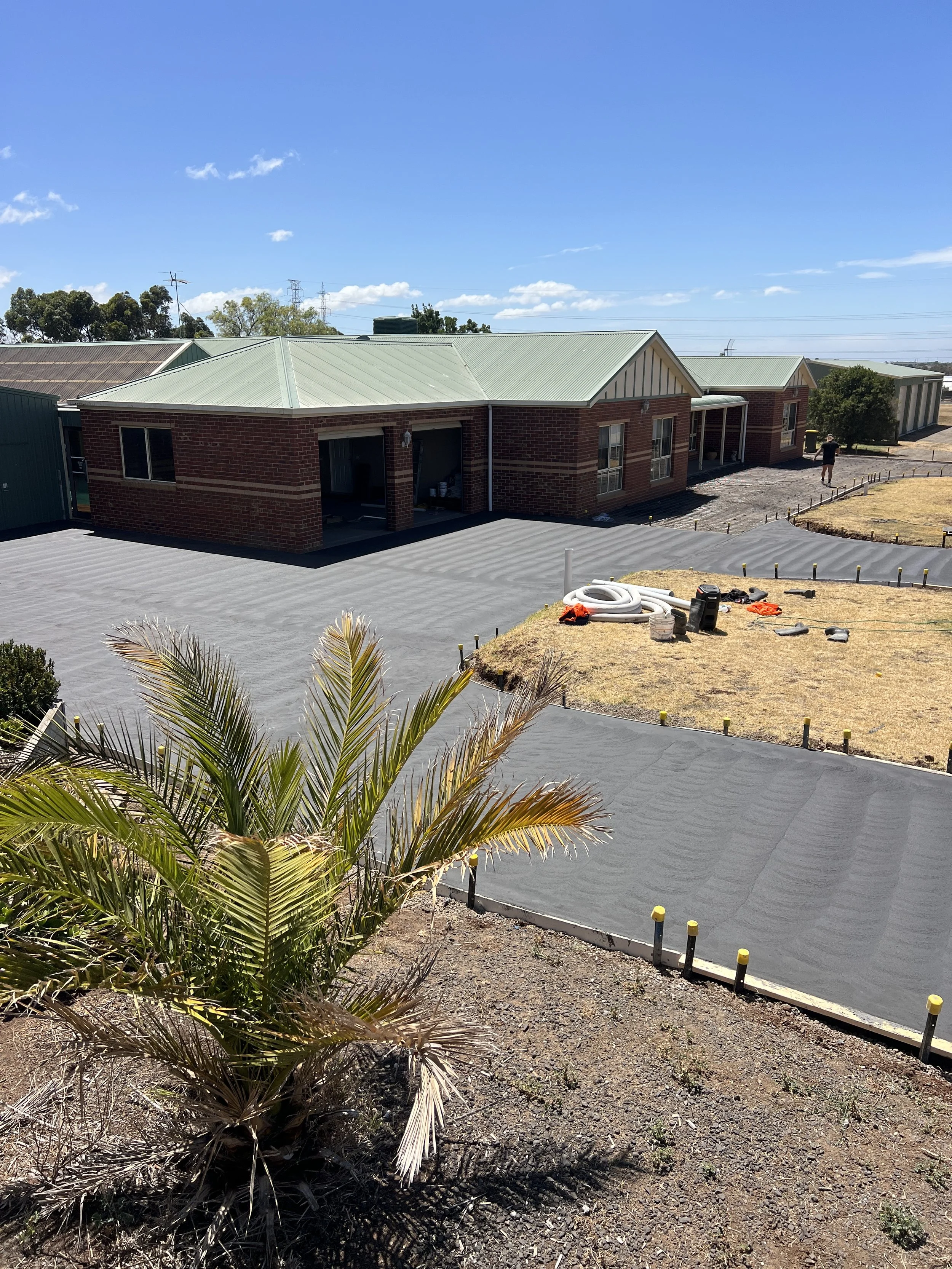 House under construction with a green metal roof, a driveway, and a person walking nearby. Construction materials and equipment are scattered around, with a palm tree in the foreground and a clear blue sky.