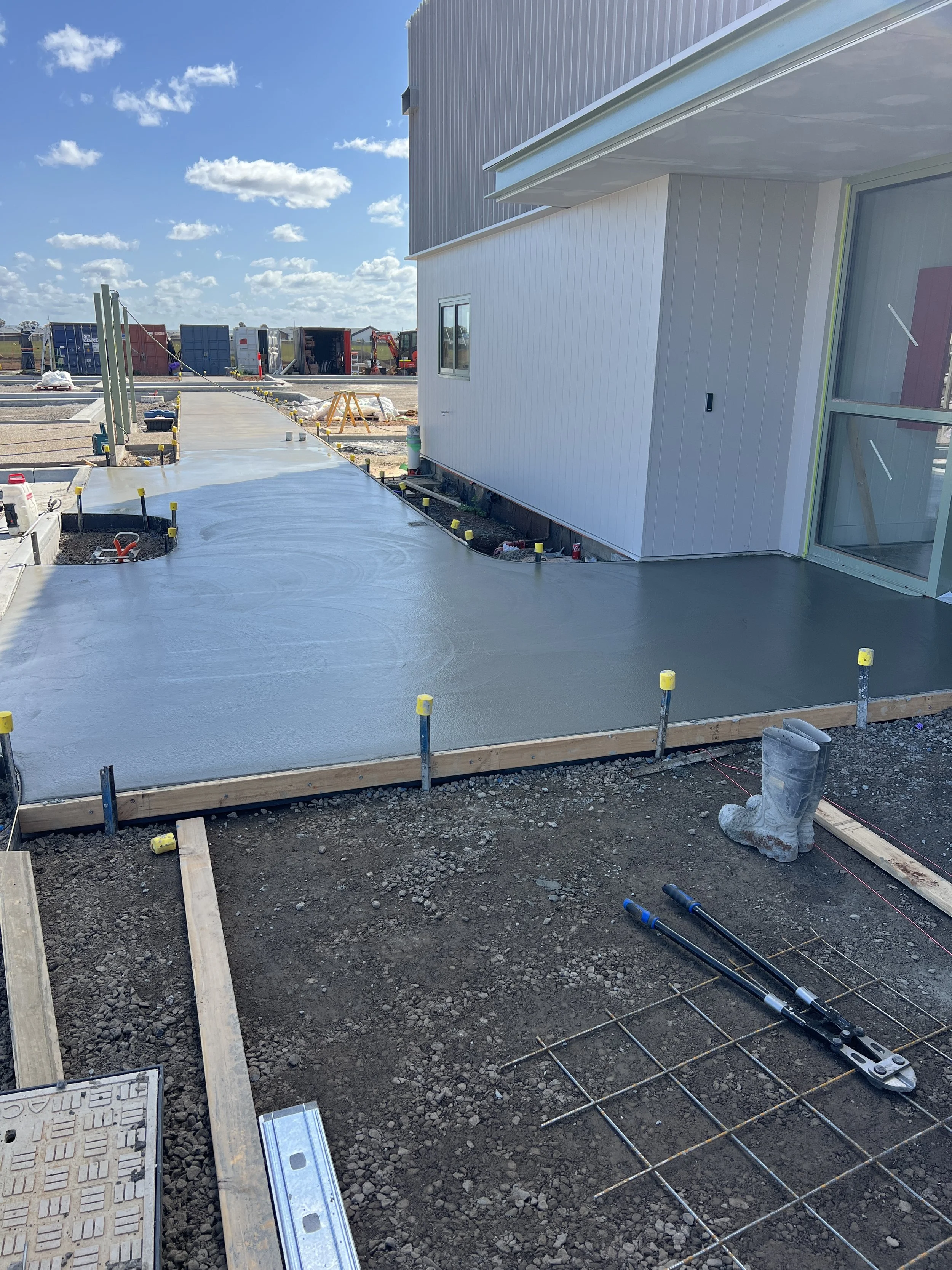 Construction site with freshly poured concrete around a building with panel siding, construction tools, and machinery in the background under a partly cloudy sky.