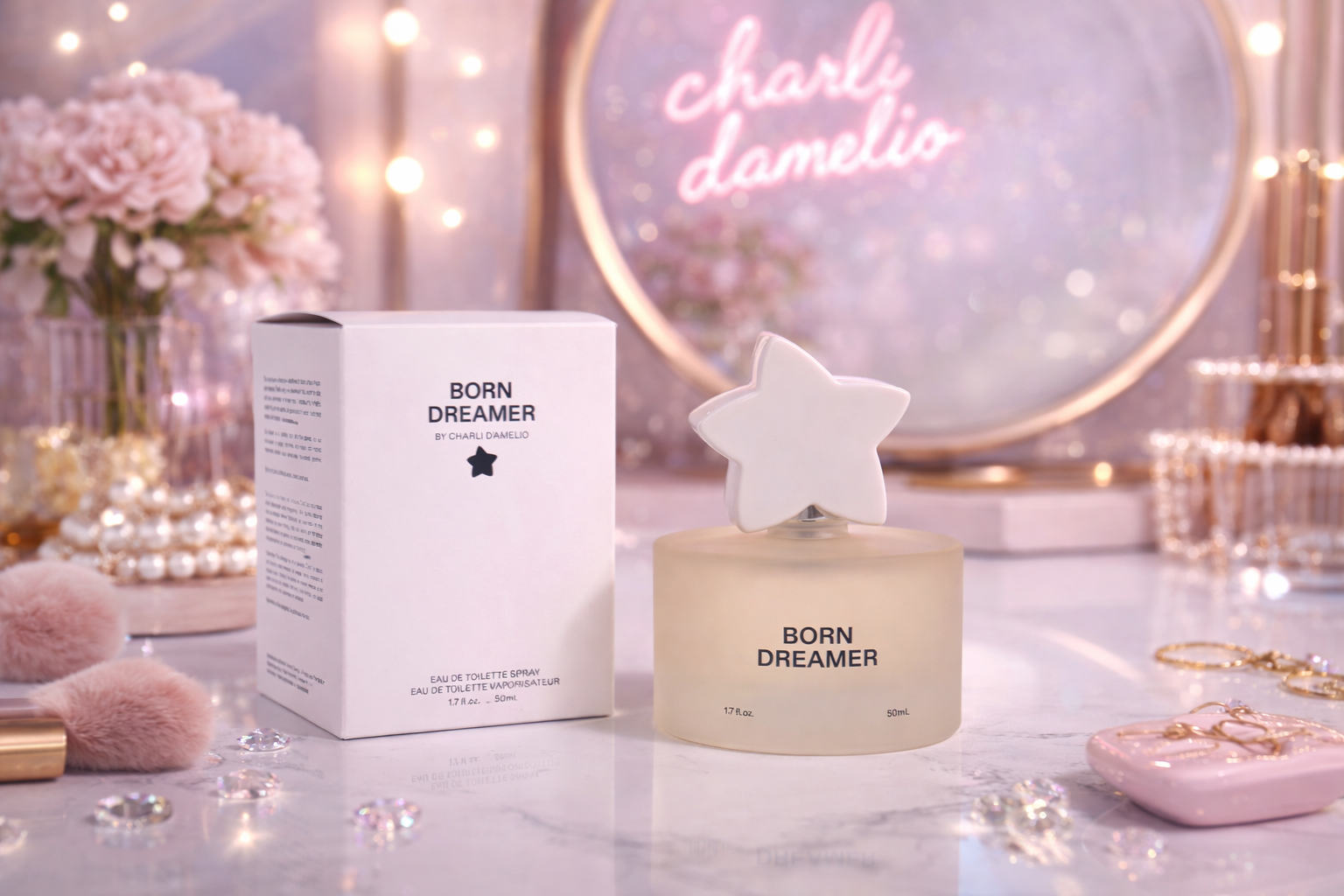 A perfume bottle labeled 'Born Dreamer' with a star on it, next to a box of the same perfume, on a vanity with pink makeup brushes, jewelry, and decorative items. In the background, a neon sign says 'charli damelio' and a mirror with pink lights surr