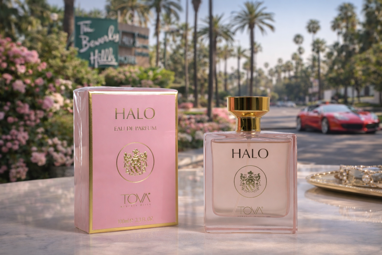 A pink perfume box labeled HALO by TOVA and a clear glass perfume bottle with a gold cap, both placed on a white marble surface outdoors with pink flowers, palm trees, and a street with cars in the background.