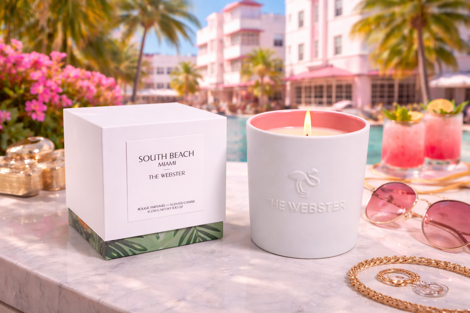 A lit scented candle in a white ceramic holder with a flaming wick, featuring an embossed flamingo and the text "The Webster." Next to it, there's a white box labeled "South Beach Miami The Webster" containing a scented candle. The background shows a