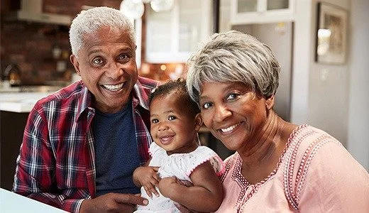 Three people, an older man, a young girl, and an older woman, smiling together indoors. The man has gray hair and is wearing a red plaid shirt, the woman has short gray hair and is wearing a light pink top, and the girl is wearing a white dress.