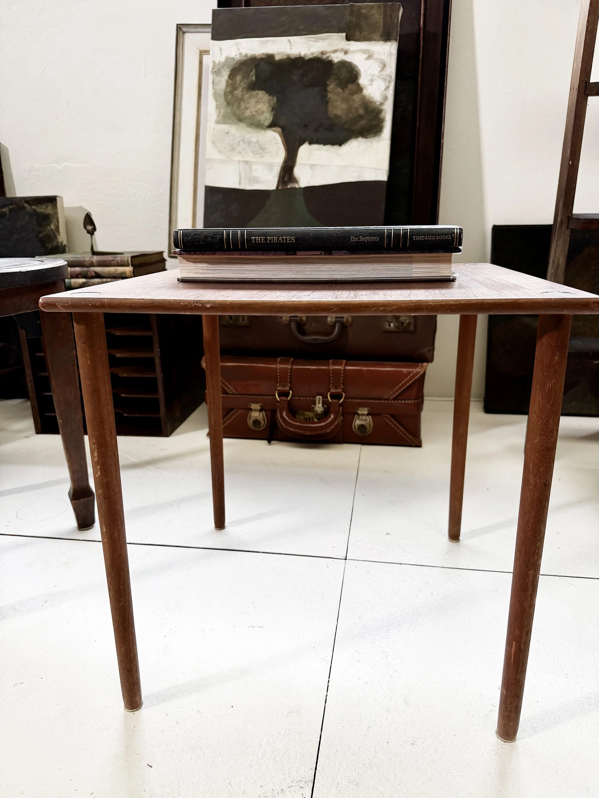 A wooden table in an Sydney art studio, there are paintings and a brown leather vintage suitcase underneath the table. The background shows a white wall with various framed artworks.