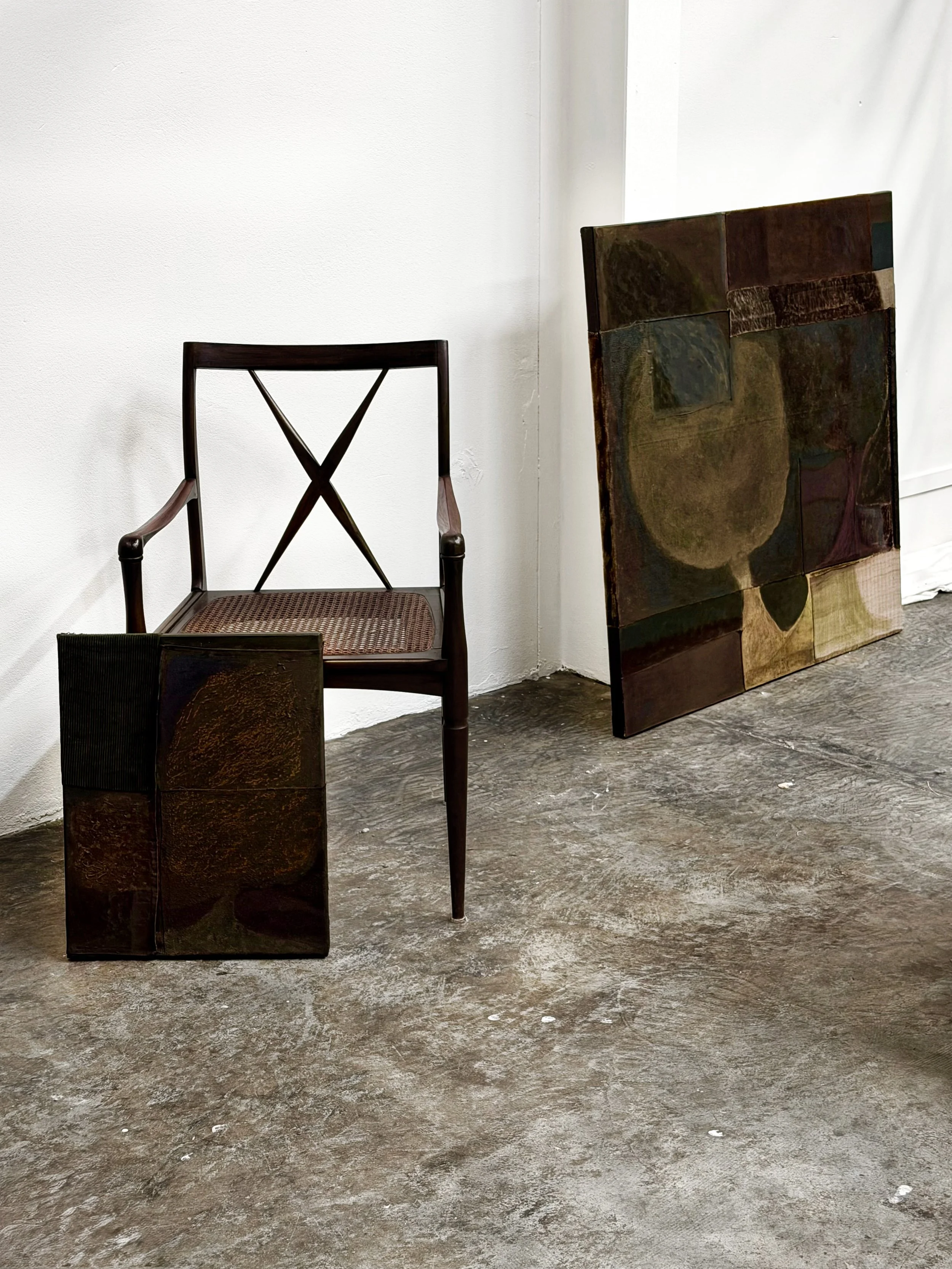 An art gallery or studio space with a wooden chair with a woven seat, two small wooden sculptures, and a large abstract painting leaning against the wall. The floor is made of concrete and the wall is plain white.