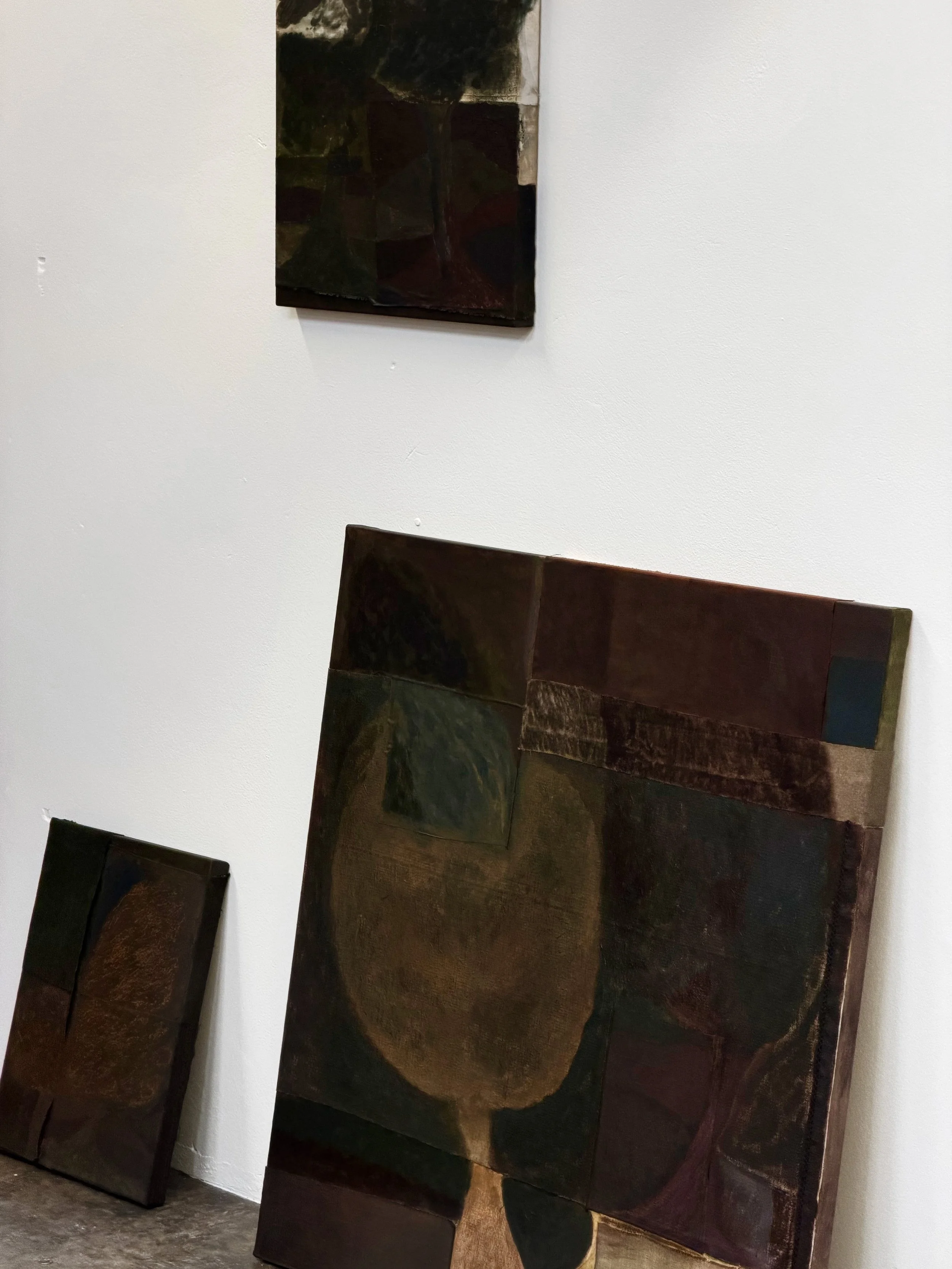 Dark abstract paintings mounted on a white gallery wall, with some artwork leaning on the floor.