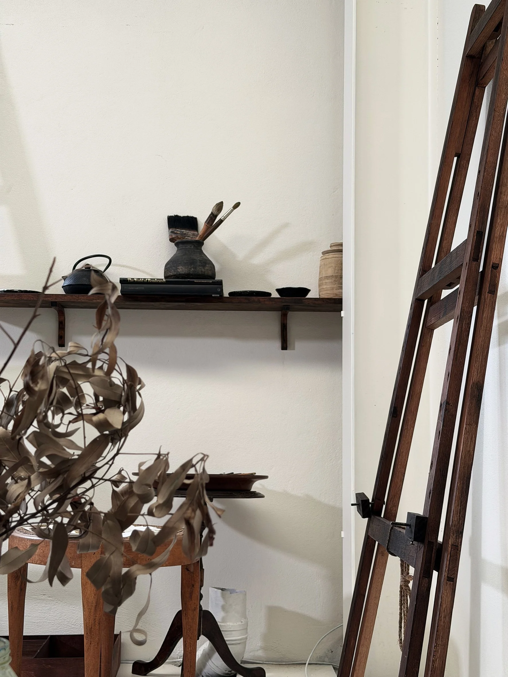 A wooden artist's easel in a sydney artspace, with a wooden table in front holding dried leaves and art supplies, including brushes and a small sculpture. There is a large ceramic pot behind the table.
