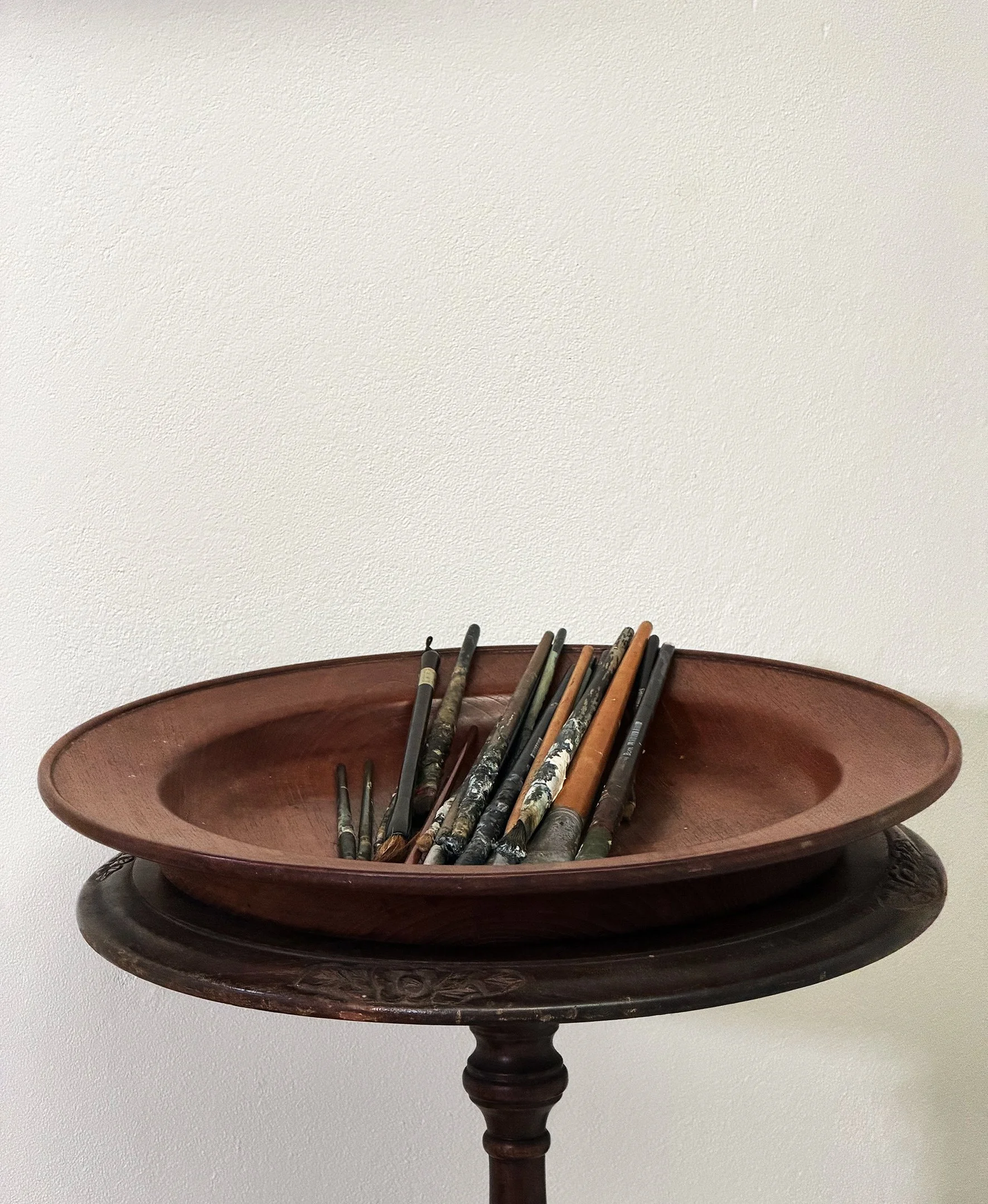 A round wooden table with a carved pedestal base, holding a large, shallow wooden bowl filled with a collection of paintbrushes and art supplies against a plain white wall background.