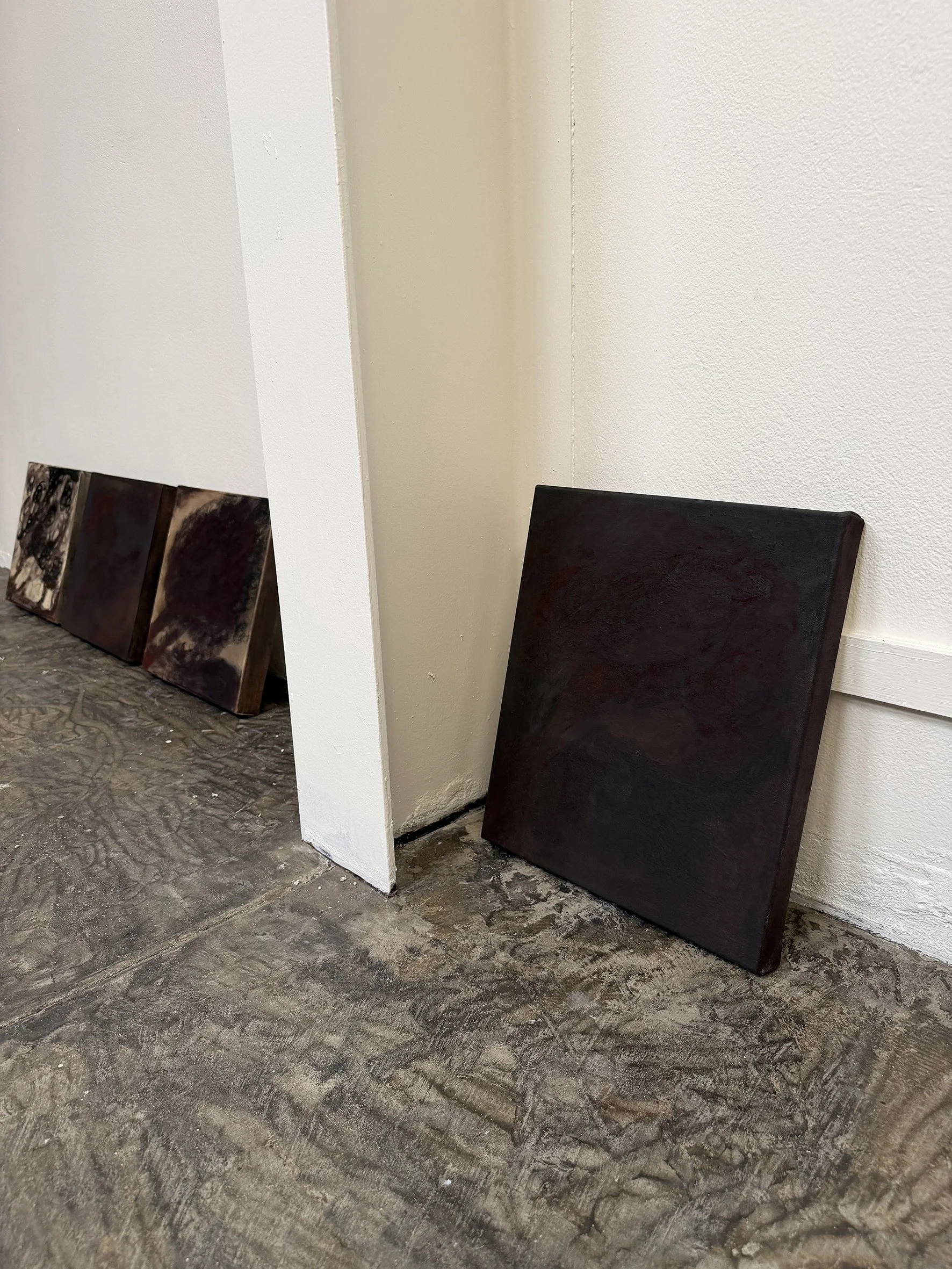 Three dark-colored abstract paintings leaning against a white wall in an art gallery, with a textured gray floor.