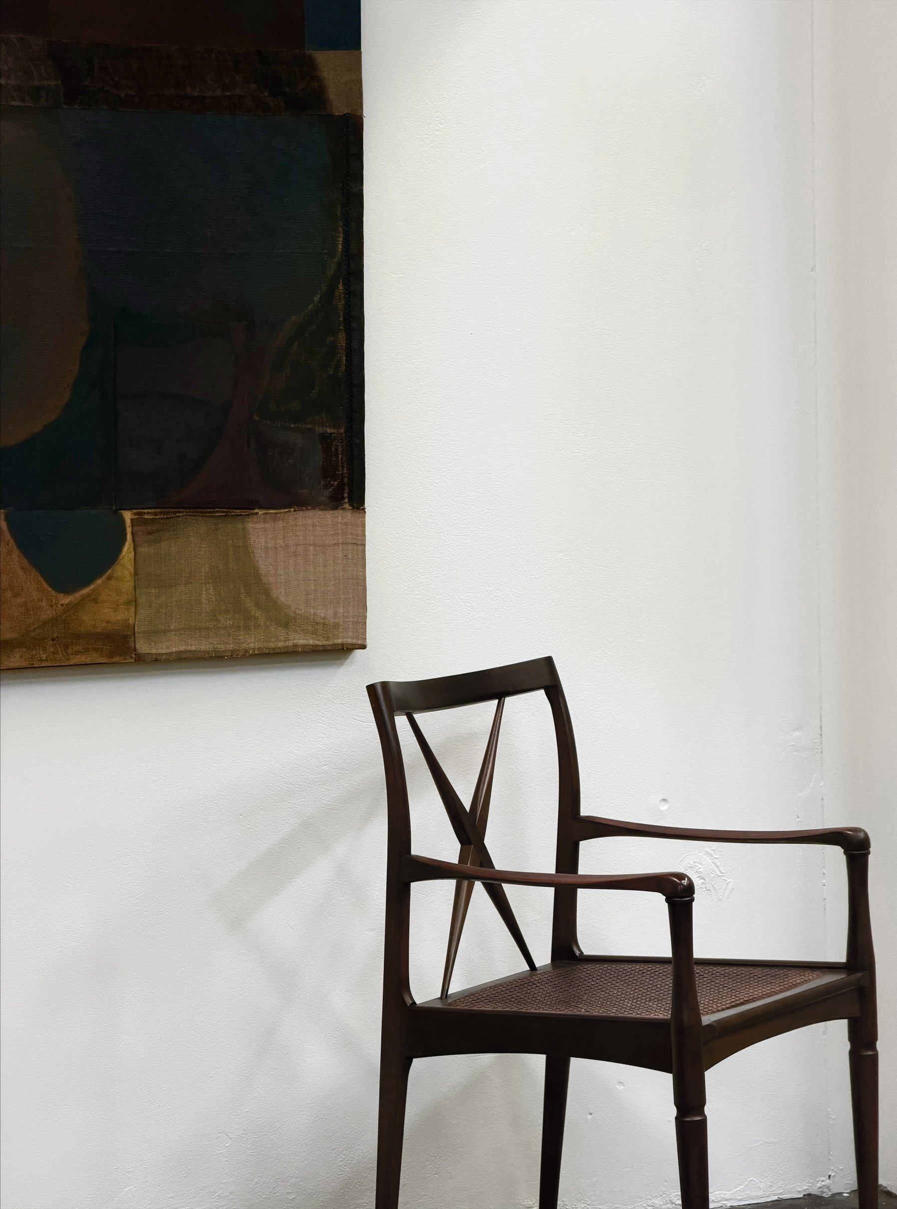 Image of a dark wooden chair with a woven seat and a decorative backrest, positioned against a plain white wall with part of a large abstract painting visible on the wall.